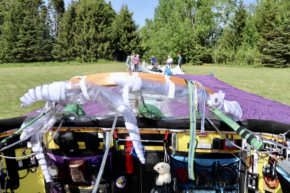A balloon gondola as it rests in a grassy field. In the distance, a group of people prepare to inflate the gas cell.