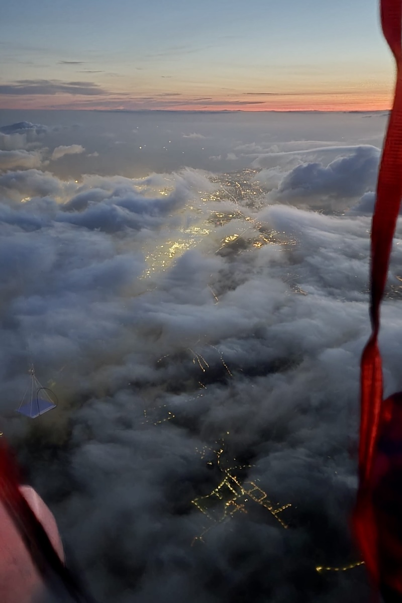 The view from a balloon gondola in the early evening sunset. The streetlights of a town glow through a thin layer of cloud.