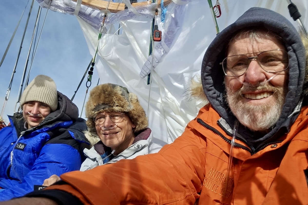 Two men and a woman dressed in heavy, winter clothing stand in a balloon gondola while it is in flight on a sunny day.