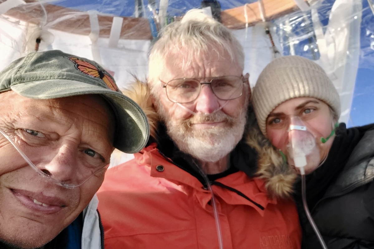 Two men and a woman dressed in heavy, winter clothing stand in a balloon gondola while it is in flight on a sunny day.