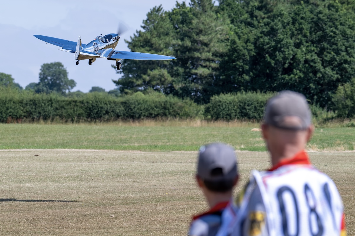A radio-controlled model of a polished aluminum Supermarine Spitfire flies away from the camera; the ground-based pilot and helper are visible in the foreground.