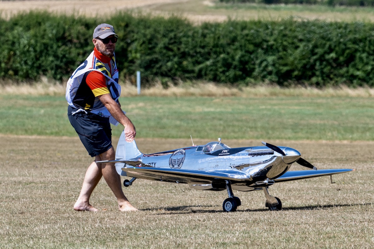A radio-controlled model of a polished aluminum Spitfire is held by its rudder while on the grass runway by a man wearing a competition bib.