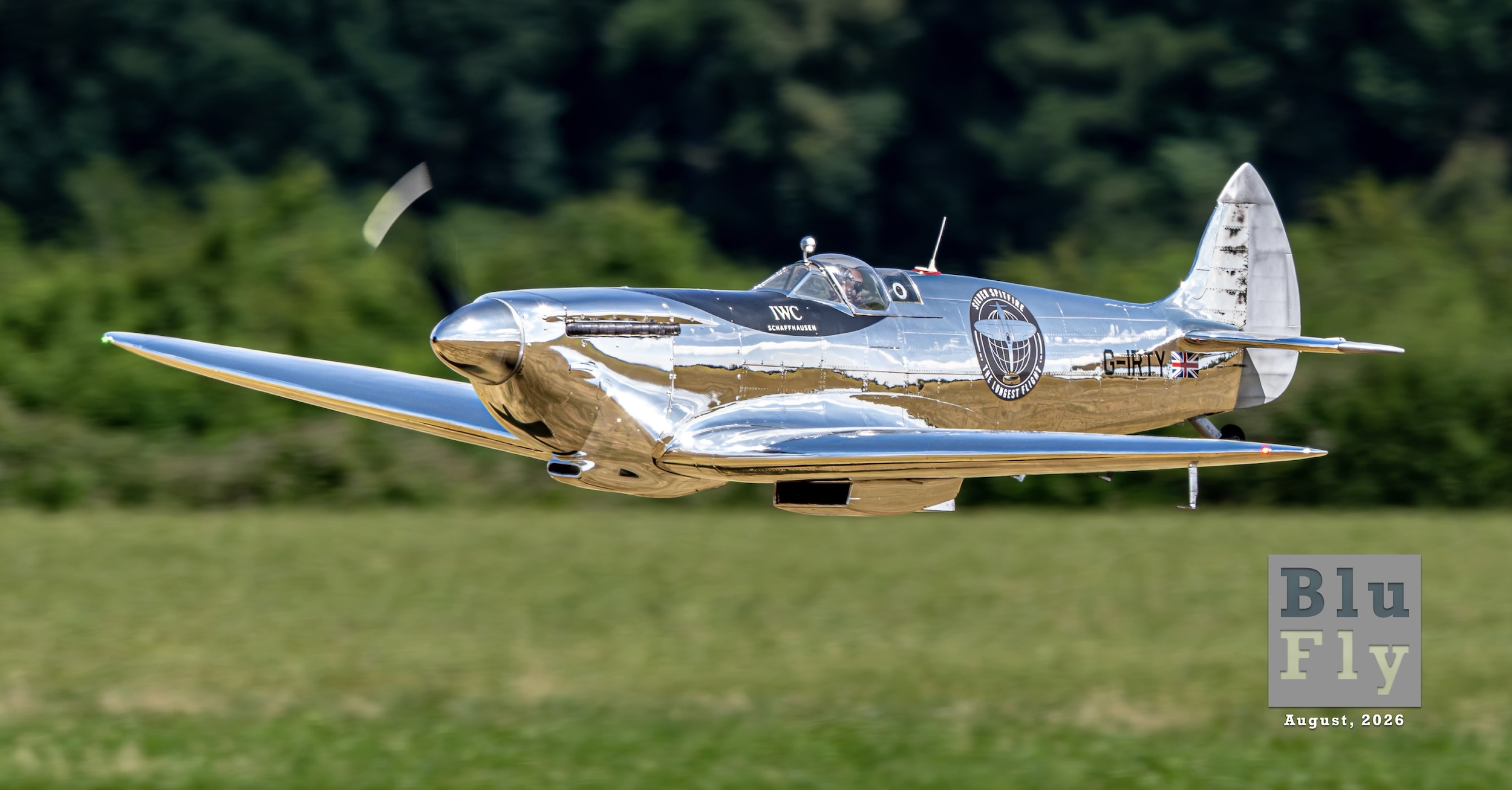 A radio-controlled model of a polished aluminum Supermarine Spitfire flies toward and to the left of the camera position against a backdrop of green trees.