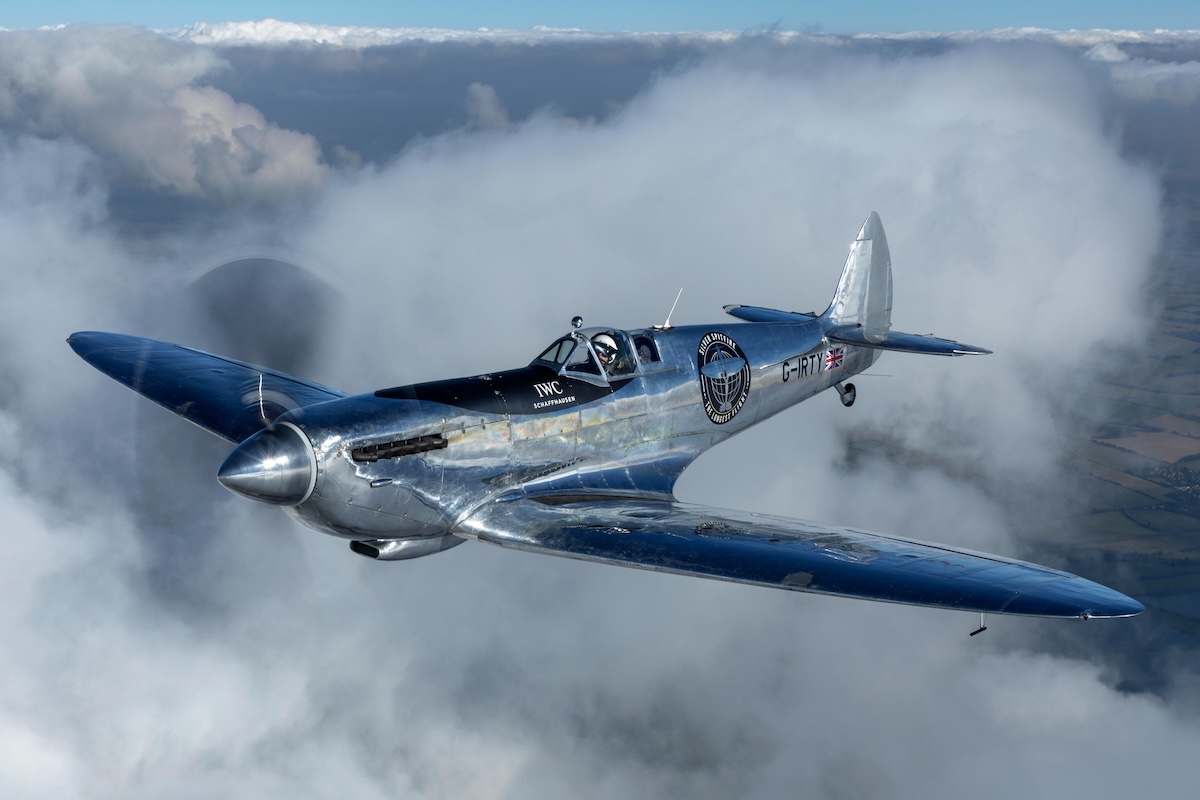 An air-to-air photo of the polished aluminum Silver Spitfire flying toward and to the left of the camera. Puffy white clouds are in the background.