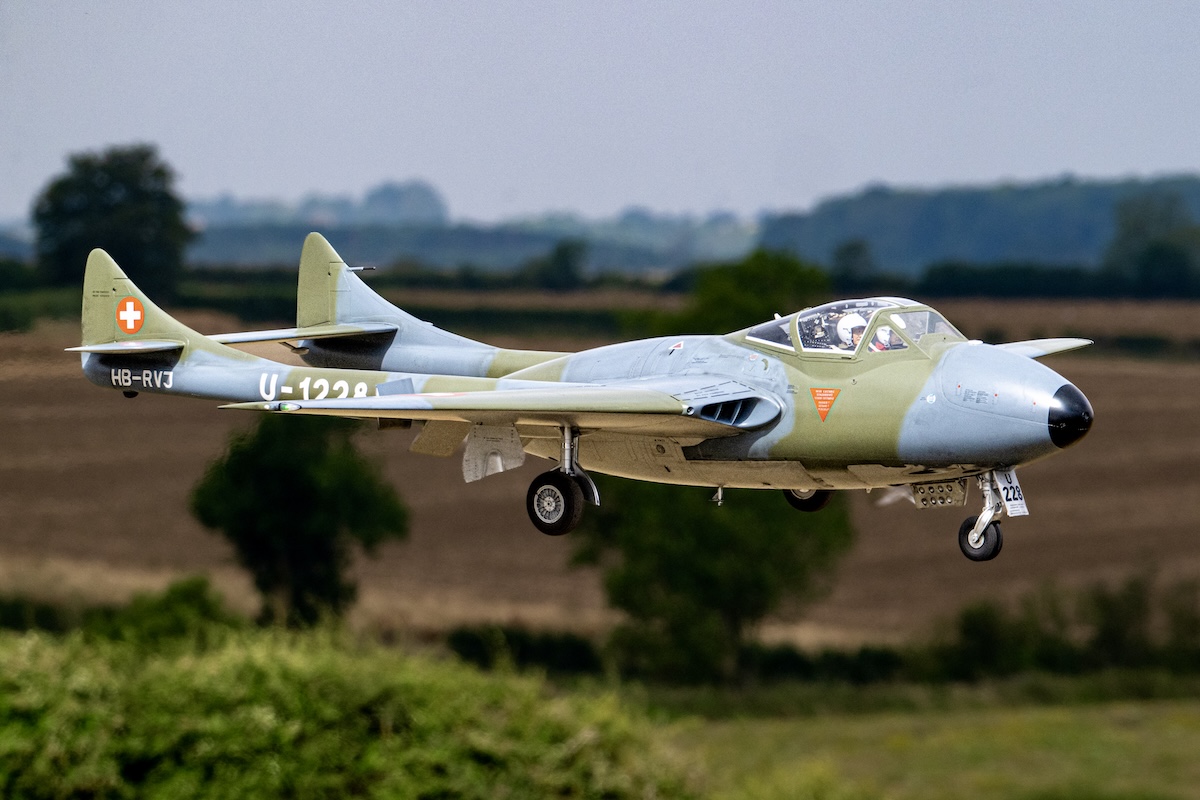 A radio-controlled model De Havilland Vampire with Swiss Air Force markings flies past the camera position. It is difficult to distinguish from the real thing.