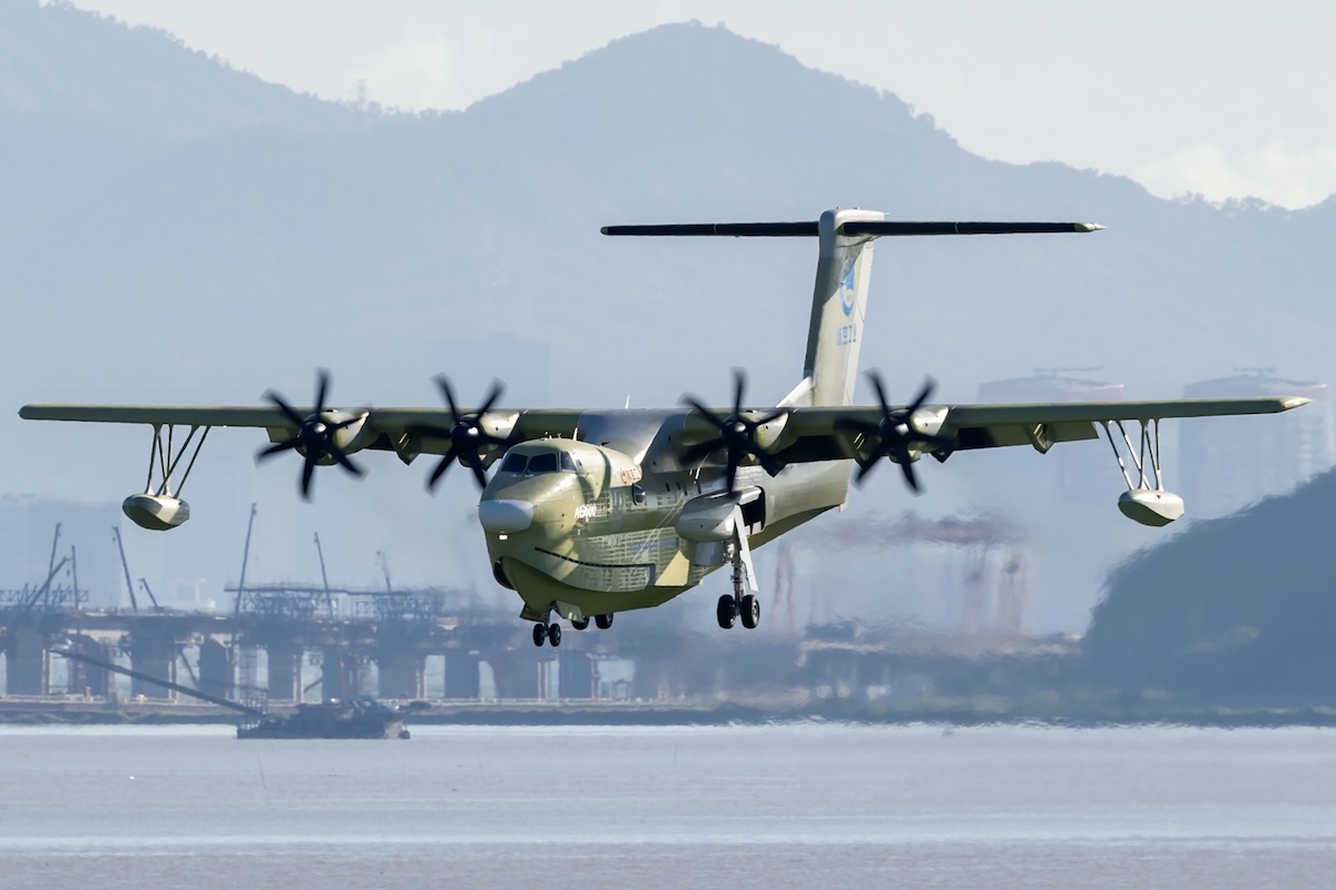 A four-turboprop AG600M flies toward the camera on what appears to be final approach. A body of water and a harbour are in the background.