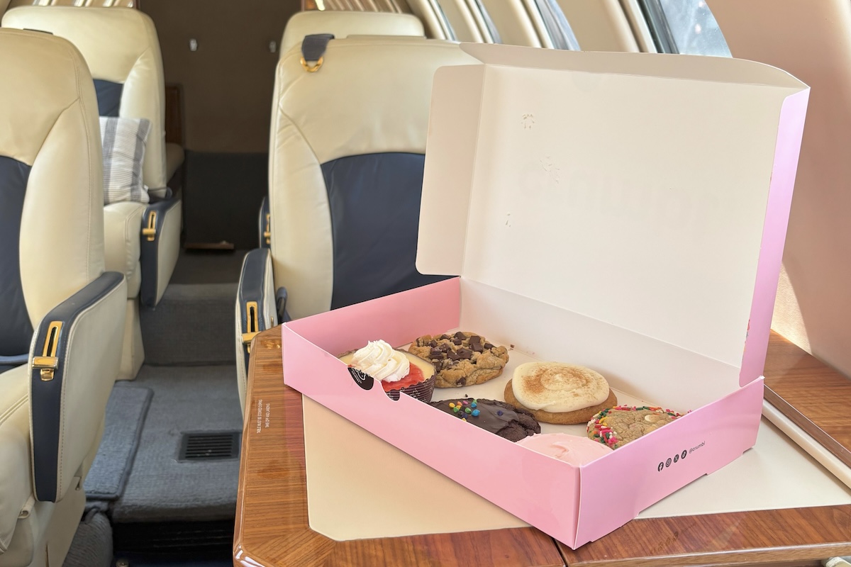 An open box of Crumbl cookies has been placed on the tray table on a seat back on a private jet.