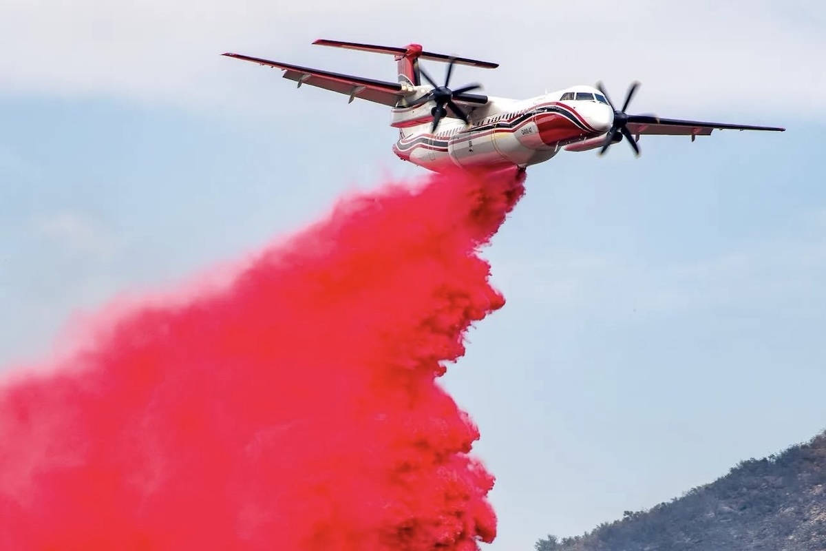 A Conair Dash 8-400AT drops its payload of red retardant set against the backdrop of a cloud-streaked sky and a tree-covered hill.