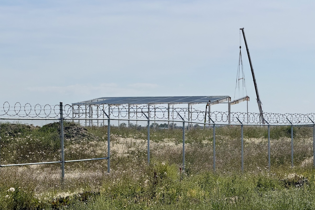 The framework of an industrial-scale hangar is visible behind a fence topped with razor wire. A construction crane is visible to the right of it.