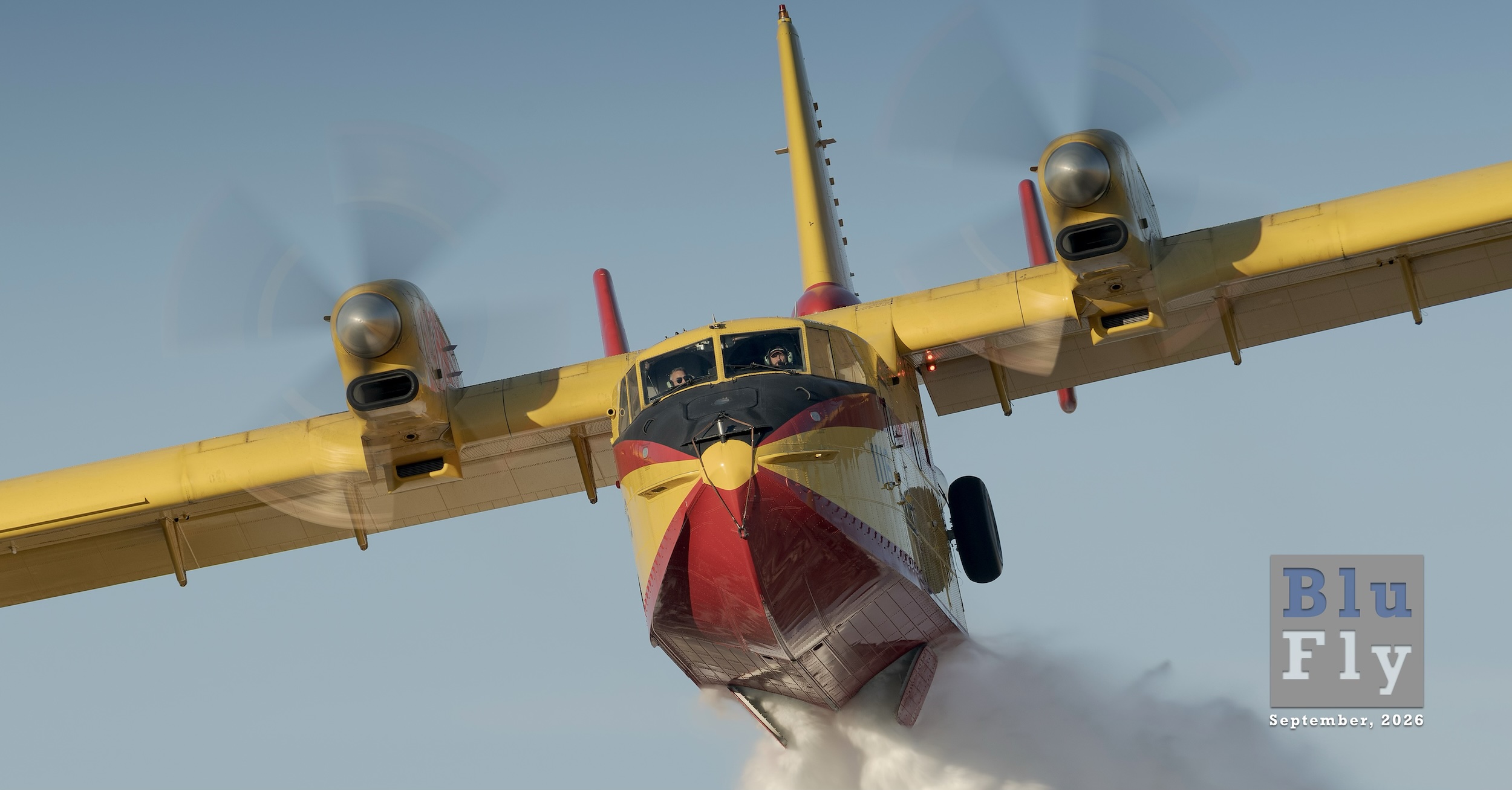 A CL-415 water bomber flies directly toward the camera position while dropping its payload of water. The pilots are visible in the cockpit.