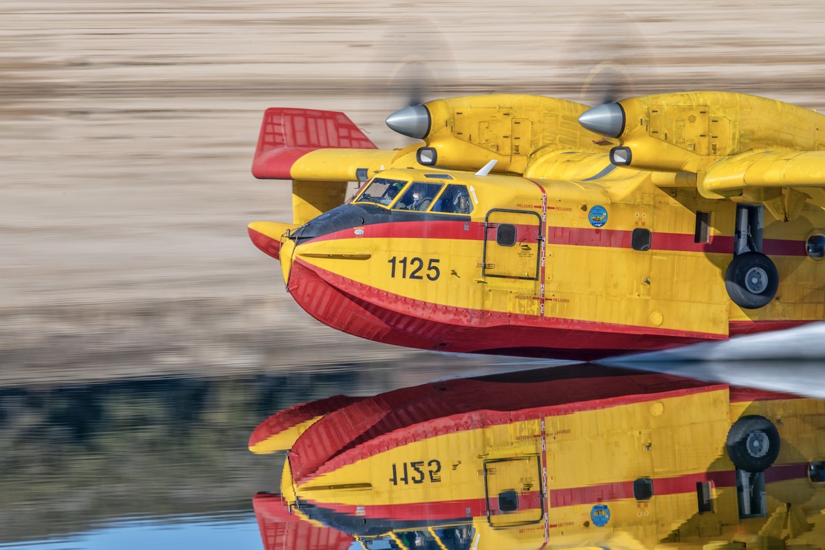 A CL-415 water bomber skims along a glassy, smooth lake so that a full reflection of the aircraft is visible.