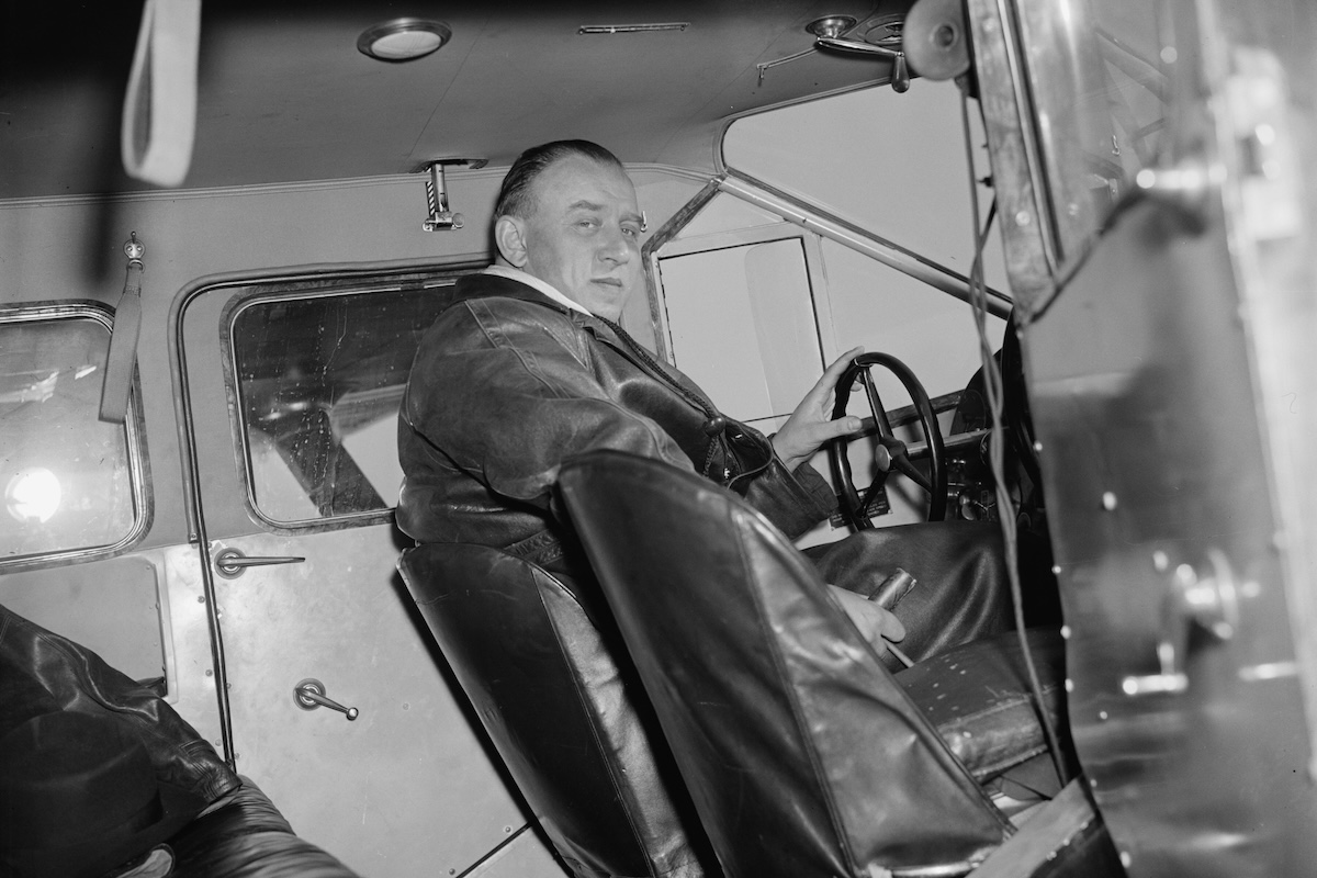 A vintage photo of a man in the left pilot seat of period-appropriate aircraft photographed through the open right door. He is smiling slightly.