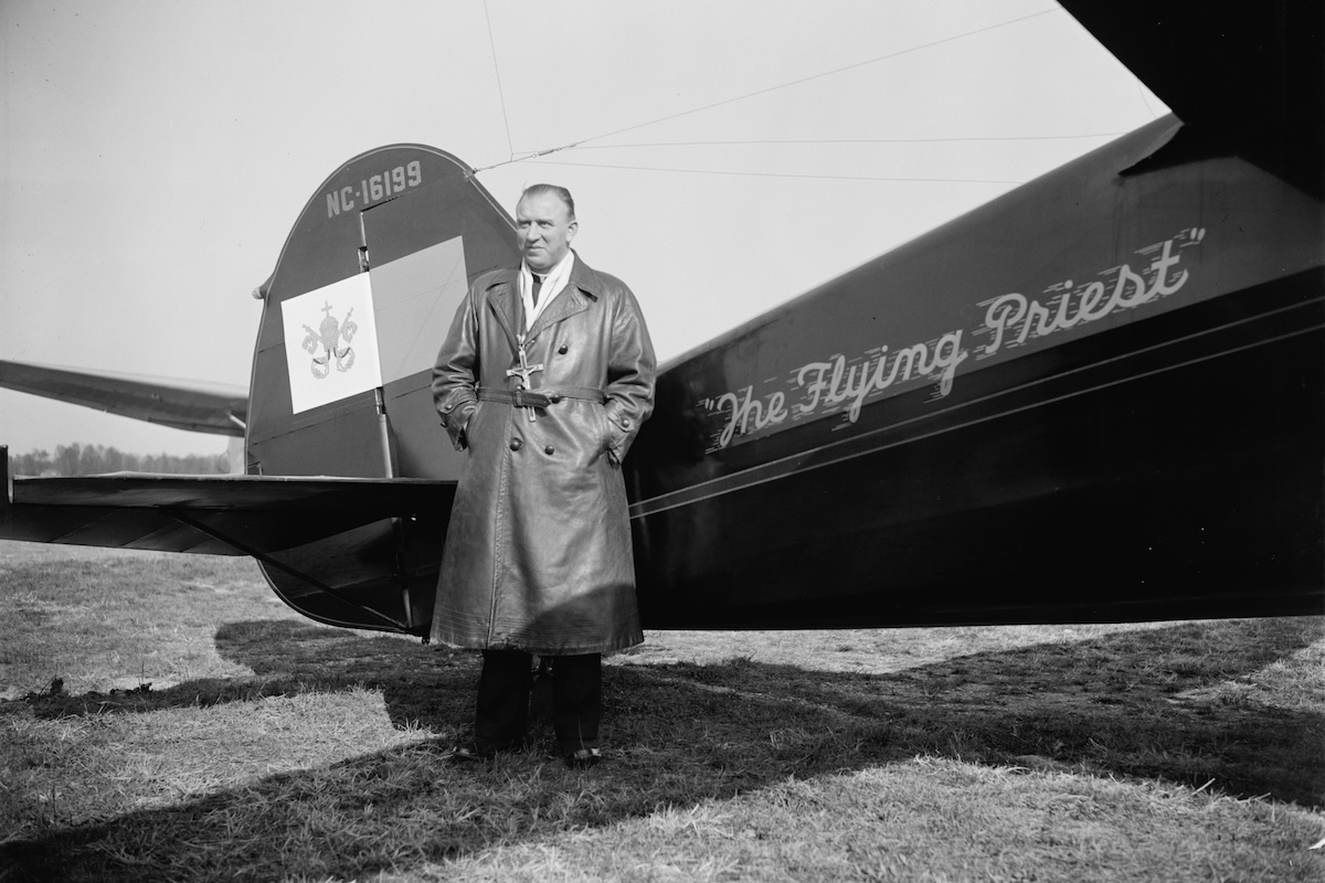 A vintage photo of a man in a long leather coat standing beside the aft fuselage of a Stinson SR-8BM Reliant aircraft, which is parked on a grass runway.