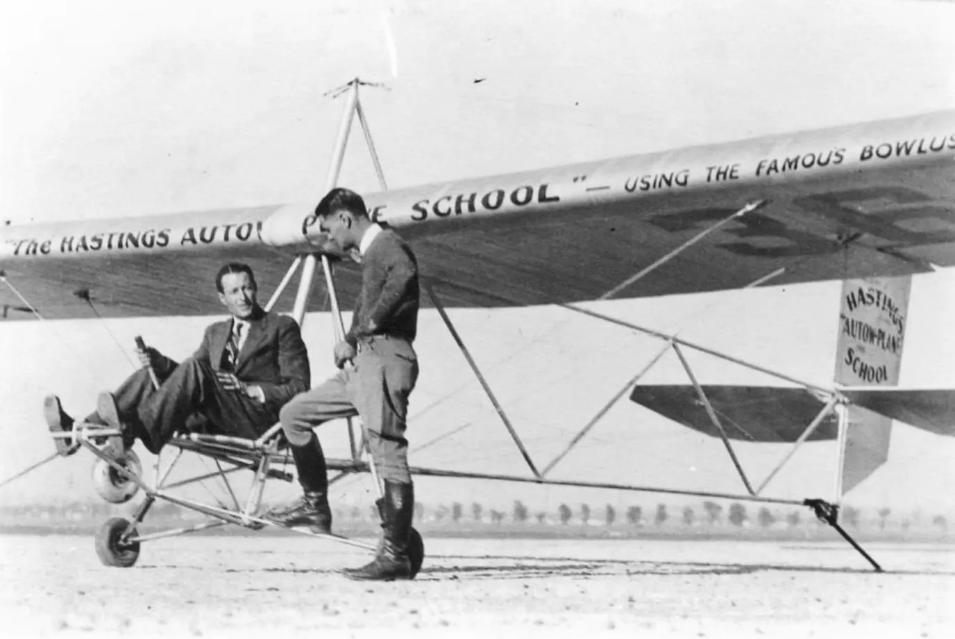 A vintage black-and-white of an old 'primary' type glider with a man seated at the controls. He is wearing a suit, light-coloured shirt, tie, and dress shoes. The glider is oriented such that it is pointed toward and to the left of the camera position, pointing to the left in the frame. A second man is standing to the left of the man seated in the pilot's seat and appears to be talking with him. He is wearing high boots, a sweater, and a white shirt. The glider's fuselage is an open framework with no external cover, which will completely expose the pilot to the airflow when the aircraft is in motion. The wing and empennage are more conventional in nature. The wing is braced with wires both above and below the wing. Painted on the leading edge of the wing are the words « THE HASTINGS AUTOW-PLANE SCHOOL » followed by, in slightly smaller letters, the words « USING THE FAMOUS BOWLUS ». Similar words are painted on the vertical stabiliser. The ground on which the aircraft is located appears to be sand or gravel. A line of evenly spaced trees is visible in the distance. The sky above is monochrome, featureless grey. Original caption from source: « Al Hastings seated in the Bowlus primary glider at Los Angeles. » (📸 ©2025 National Soaring Museum, all rights reserved.)