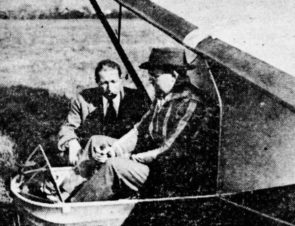 A grainy, vintage, black-and-white photo of a man sitting in a glider. The man is wearing a chequered shirt and fedora. He has both hands on the control stick. The glider is oriented such that it is pointing to the left in the frame and is canted to the left, presumably from having its left wing touching the ground. The fuselage consists of an open frame and is of high-wing, 'primary' type. A second man crouches next to the glider, on the far side of the fuselage, and appears to be giving instructions to the man in the glider. The man giving the instructions is wearing a suit, white shirt, and tie. Original caption from source: « Al Hastings instructing Franklin Iszard in a Franklin PS-2 glider, perhaps the same as the one used by Hastings to win the 1930 Nationals. » (📸 Elmira Star-Gazette, October 27, 1930)
