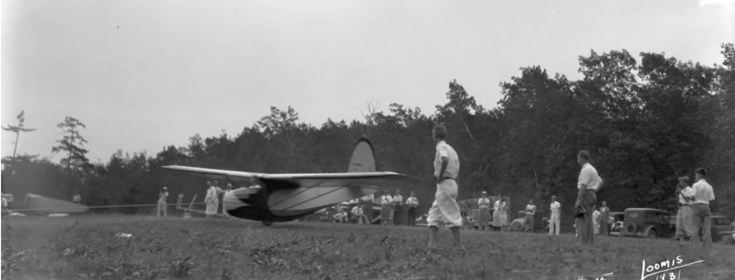 A vintage black-and-white photo showing a glider on some sort of accelerating away from a crowd of spectators on a grass runway. The aircraft is moving from right to left in the frame. There appears to be a small amount of daylight below the single-wheel landing gear on the bottom of the fuselage, indicating the glider travelling at something approaching take-off speed. The glider is of vintage type with a high wing and supporting struts, and a rounded vertical stabiliser and rudder. Three men stand adjacent to the glider in a manner indicating they assisted with the take-off. A line of trees is visible beyond the group of spectators in the background, behind the glider. The sky is featureless, monochrome grey. Original caption from source: « A Franklin PS-2 taking off by bungee cord from one of the hills near Elmira, NY. » (📸 ©2025 National Soaring Museum, all rights reserved.)