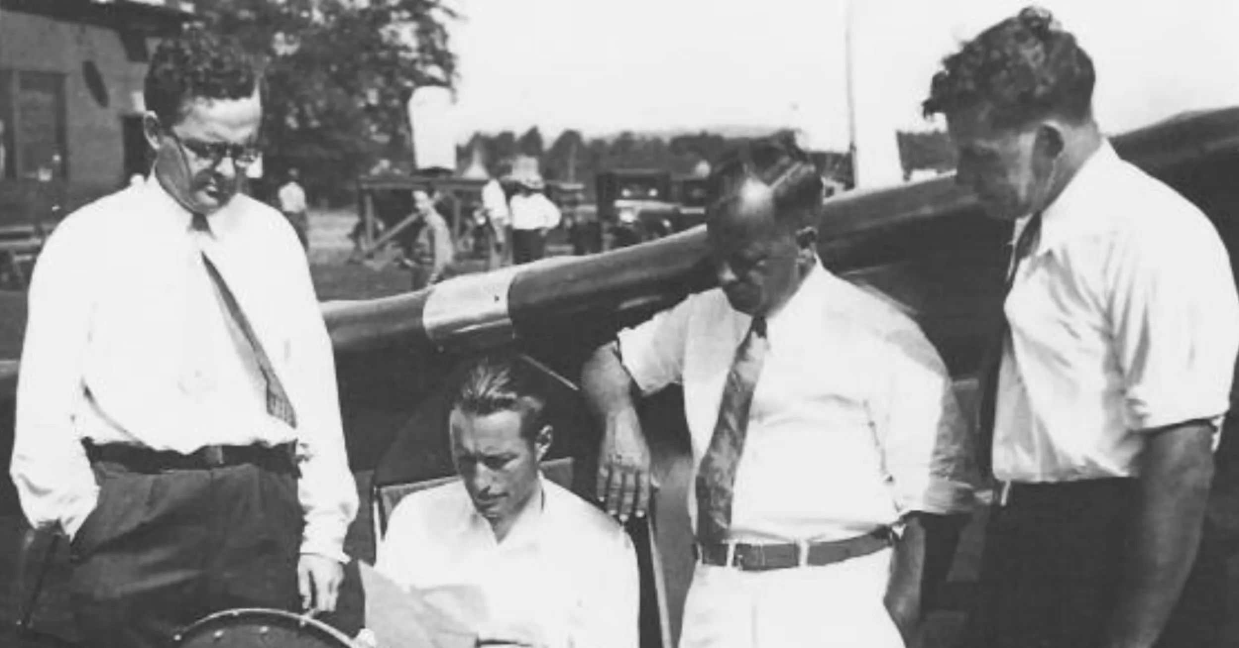 A vintage black and white photo of four men and a glider. Three of them are standing, and the fourth, second from left, is seated in the single seat of the glider itself. The three men who are standing are all wearing white shirts and ties. The man seated in the glider is also wearing a white shirt but no tie. The attention of all of the men is on a sheet of paper held by the man seated in the glider. There is activity beyond the wing of the glider in the form of people milling around, consistent with an outdoor event. Original caption from source: « ranklin 'Bud' Iszard, Al Hastings, Warren Eaton and Hawley Bowlus at the 1930 National Contest. » (📸 ©2025 National Soaring Museum, all rights reserved.)
