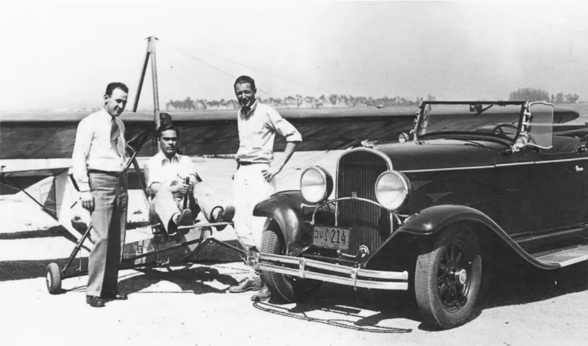 A vintage black-and-white photo of an old 'primary' type glider with a man seated at the controls. He is wearing a white, open-neck collared shirt. The glider is oriented such that it is pointed almost directly towards and to the right of the camera position, pointing to the right in the frame. To the immediate right of the glider is an old, convertible car parked pointing toward and to the left of the camera. Two men are standing next to the glider, to the left and right of the pilot. All three men are looking directly at the camera. The man on the left of the fuselage is wearing a white shirt and tie. The man on the right is also wearing a white, open-neck collared shirt. The pilot has both hands on the glider and his feet on the rudder bar, presumably used for rudder control. The glider's fuselage is an open framework with no external cover, which will completely expose the pilot to the airflow when the aircraft is in motion. The wing and empennage are more conventional in nature. The wing is braced with wires both above and below the wing. The ground on which the aircraft is located appears to be sand or gravel. A line of evenly spaced trees is visible in the distance. The sky above is monochrome, featureless grey. Original caption from source: « Primary glider instruction at Mines Field, Los Angeles with Hastings shown standing on the right. » (📸 ©2025 National Soaring Museum, all rights reserved.)