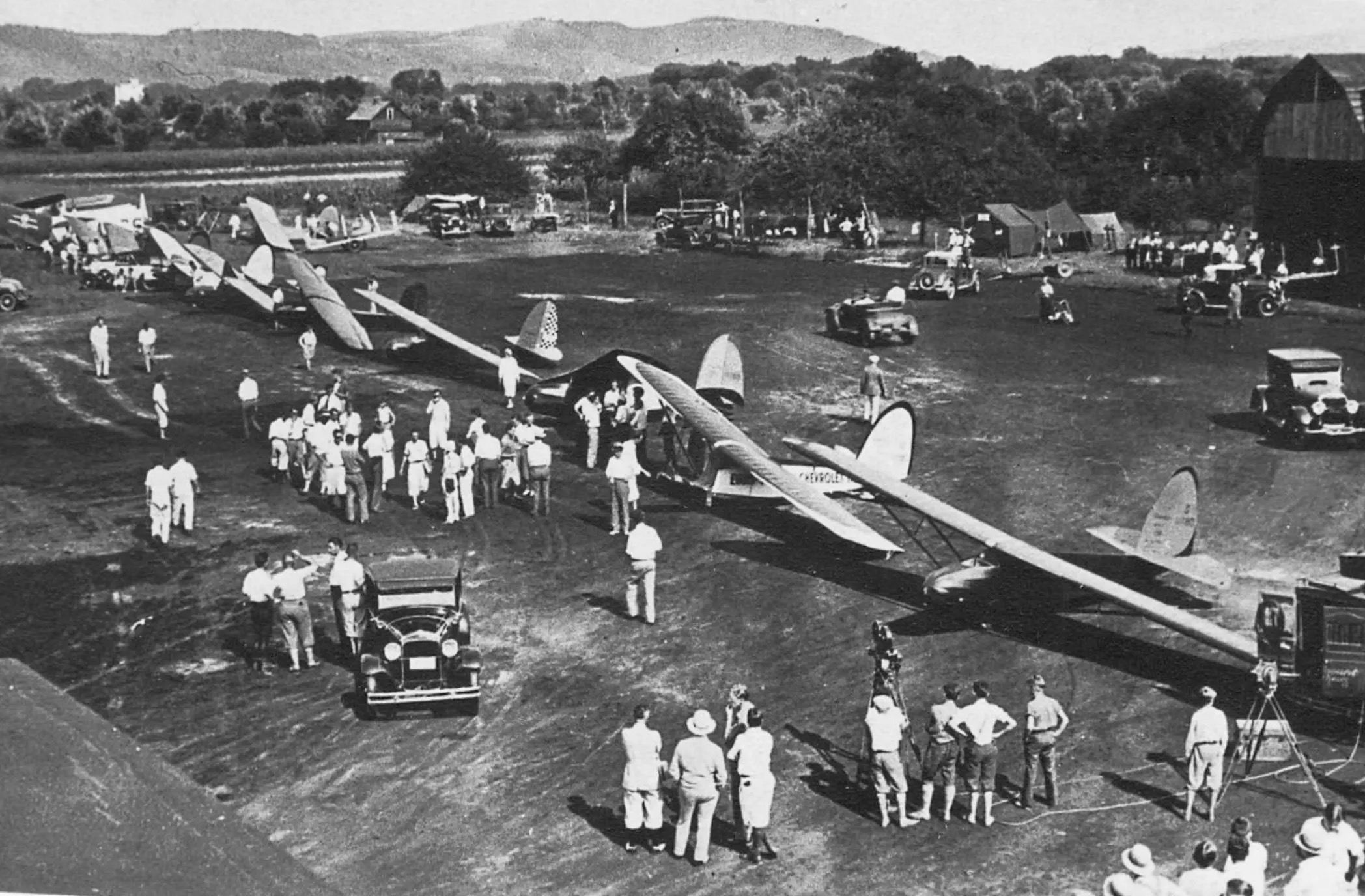 A vintage, black-and-white image, taken from an elevated position, of a glider meet taking place in a clearing surrounded by trees and agricultural fields. There are a dozen or so vintage gliders lined up seemingly ready to be launched. A significant number of people and old cars are intermingled with the gliders. A large barn is visible on the right side of the frame. Rolling hills are visible in the distance. Original caption from source: « The scene at the Second National contest 1931 at the old Caton Ave. Airport of Elmira, New York. » (📸 ©2025 National Soaring Museum, all rights reserved.)