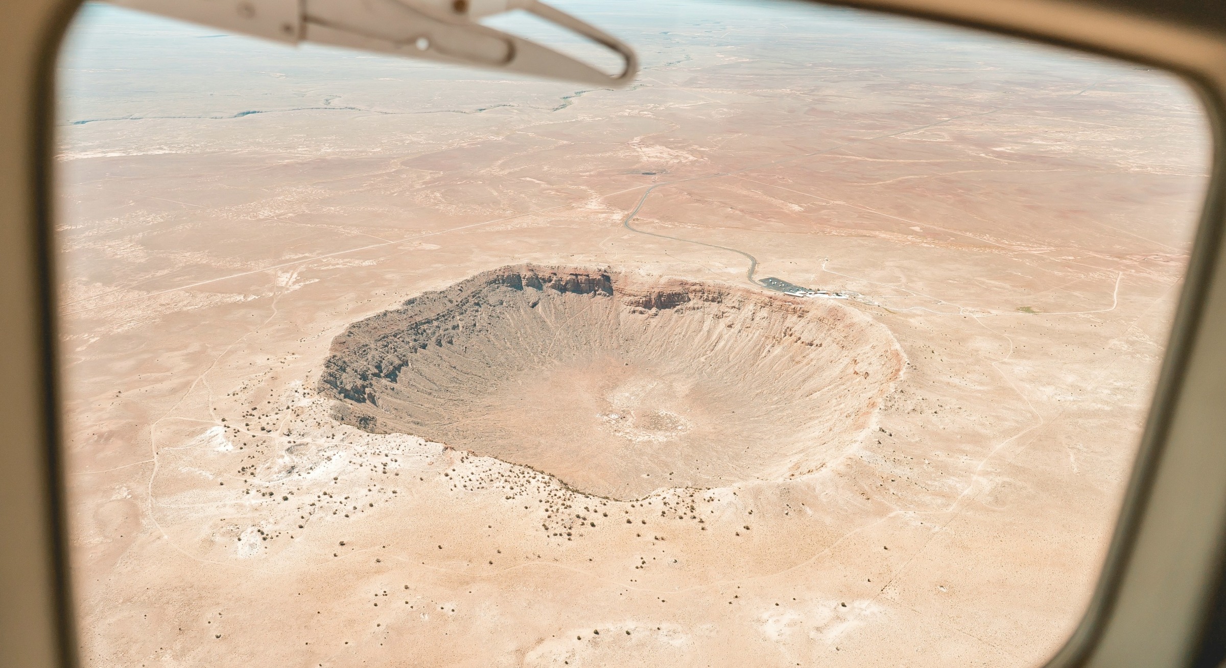 An image from an airplane window of the meteor crater located near Winslow, Arizona.  The landscape if arid and brown.  Beyond the crater it's possible to see the visitor centre as well as the road leading up to it. The sky above the desert is clear and blue with a bit of haze.