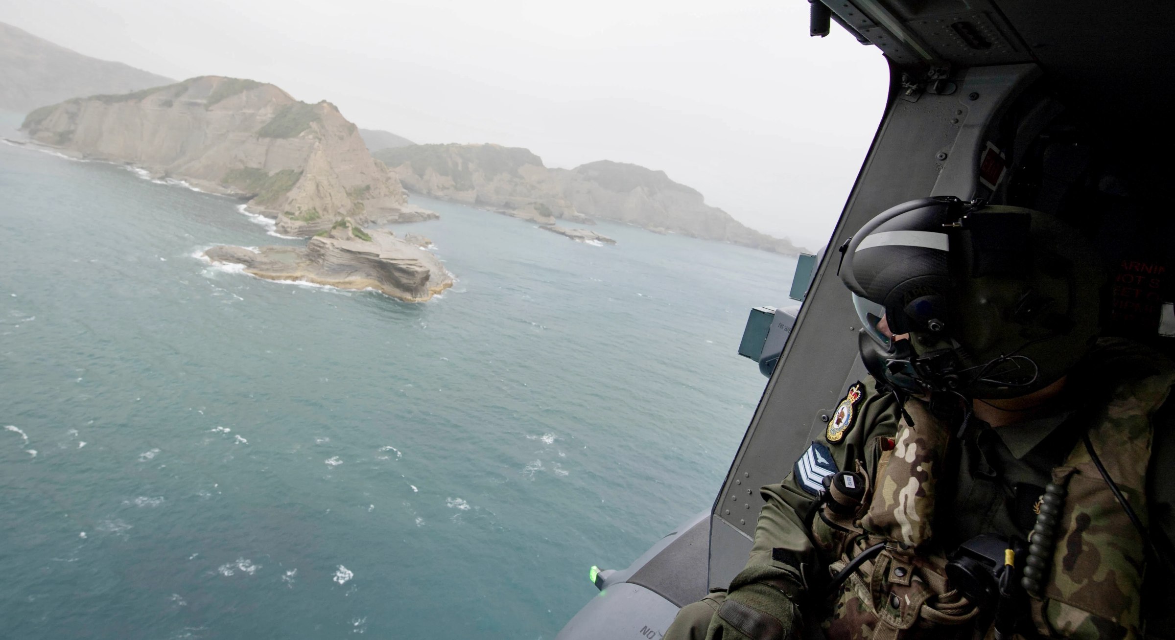 A person dressed in a military uniform and helmet sits in the open door of a helicopter as it flies low over a rocky headland. The person has their head turned such that it's not possible to see their face. The land appears to be brown and dry.  The ocean is a beautiful blue colour and whitecaps can be seen atop some of the waves. The sky is a hazy overcast tending toward blue. The original caption from the source reads: « A Helicopter Loadmaster from the Royal New Zealand Air Force’s №3 Squadron scours the sea during patrols conducted by the New Zealand Defence Force and the Ministry for Primary Industries over the east coast of the North Island. » The credit for both the photo and the caption is New Zealand Defence Force under CC BY-NC 2.0 DEED.