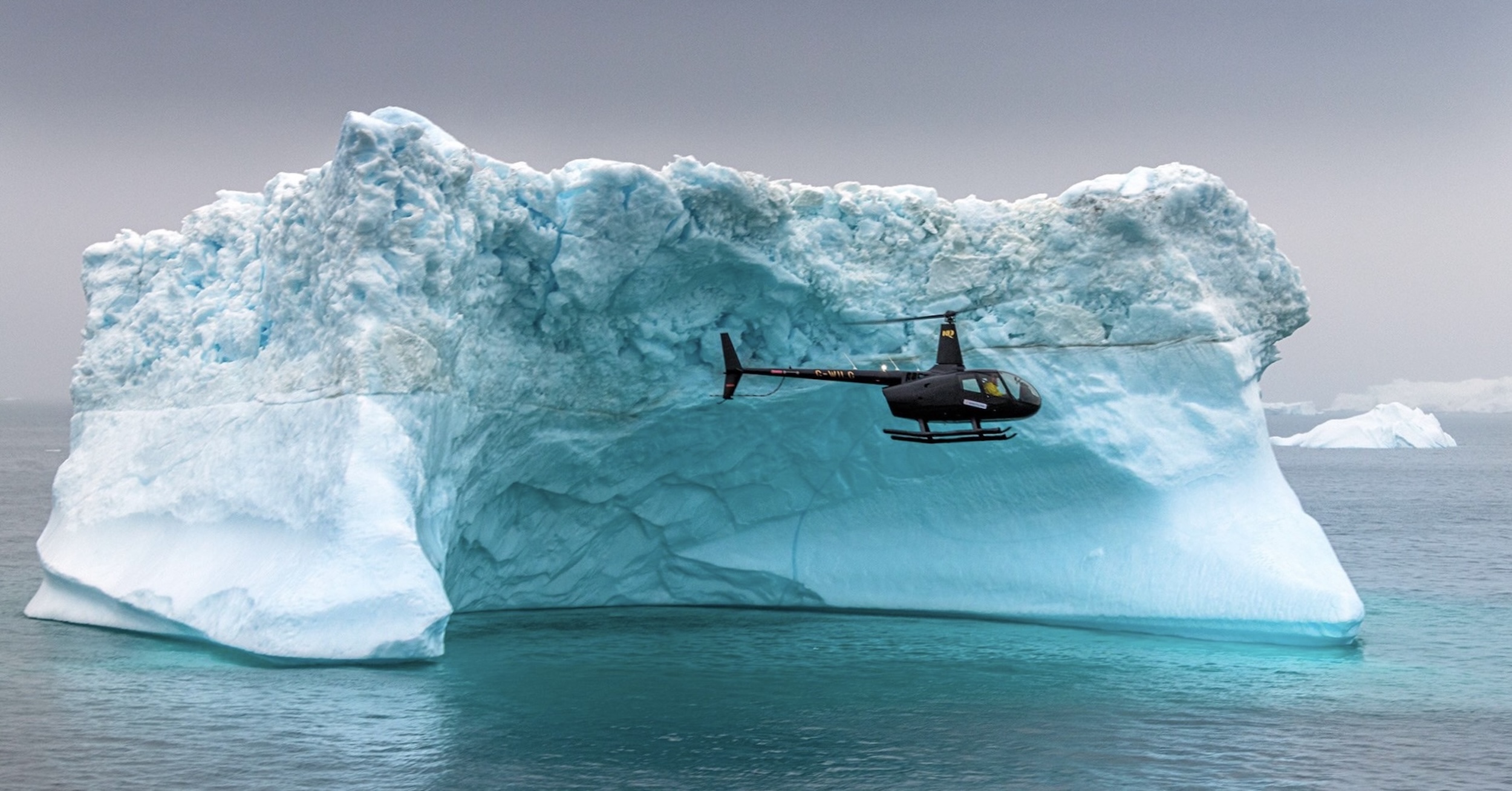A dark-coloured helicopter flies past a blue/white iceberg. In the distance, other icebergs are visible, and then a uniformly grey sky as the background.