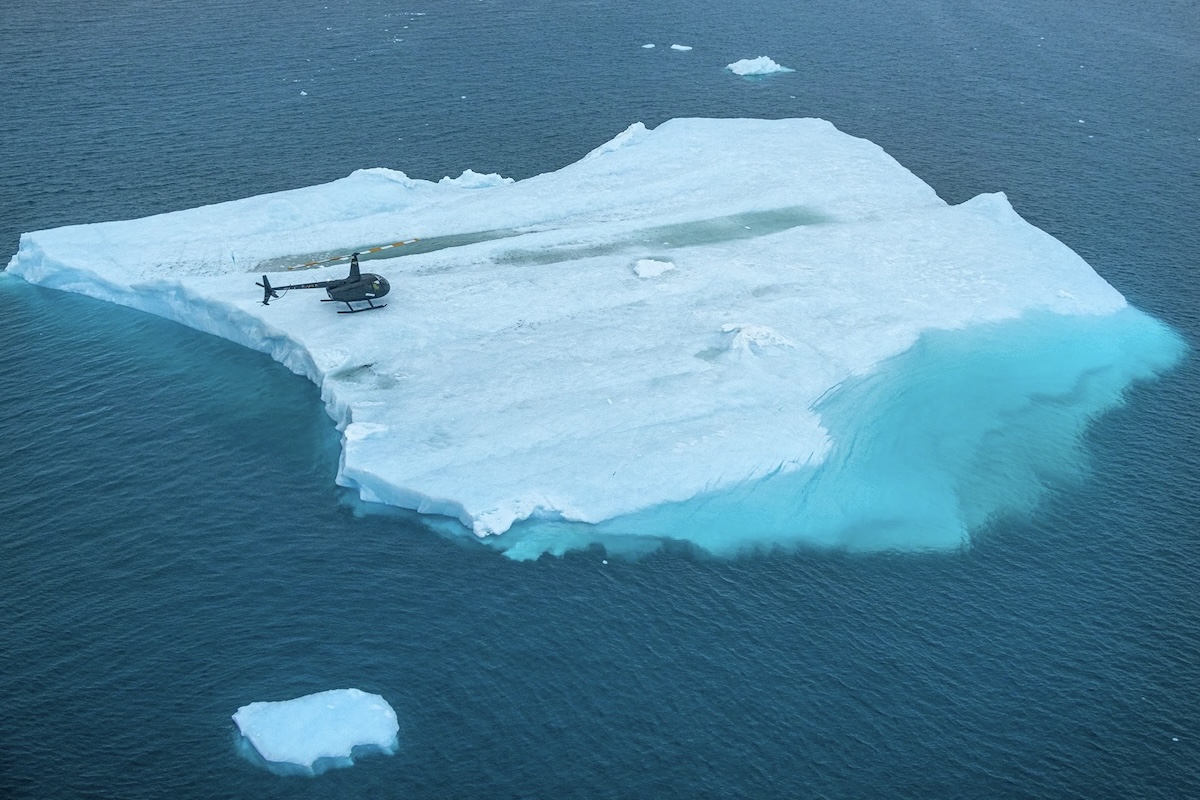 A dark-coloured helicopter with red and white blades after having landed on a flat iceberg floating in a calm blue sea. Smaller icebergs float nearby. 