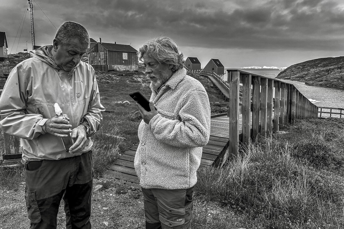 Two men stand next to an elevated, wooden walkway leading toward some small, detached, modern-looking cabins. One man is talking on a cell phone. 