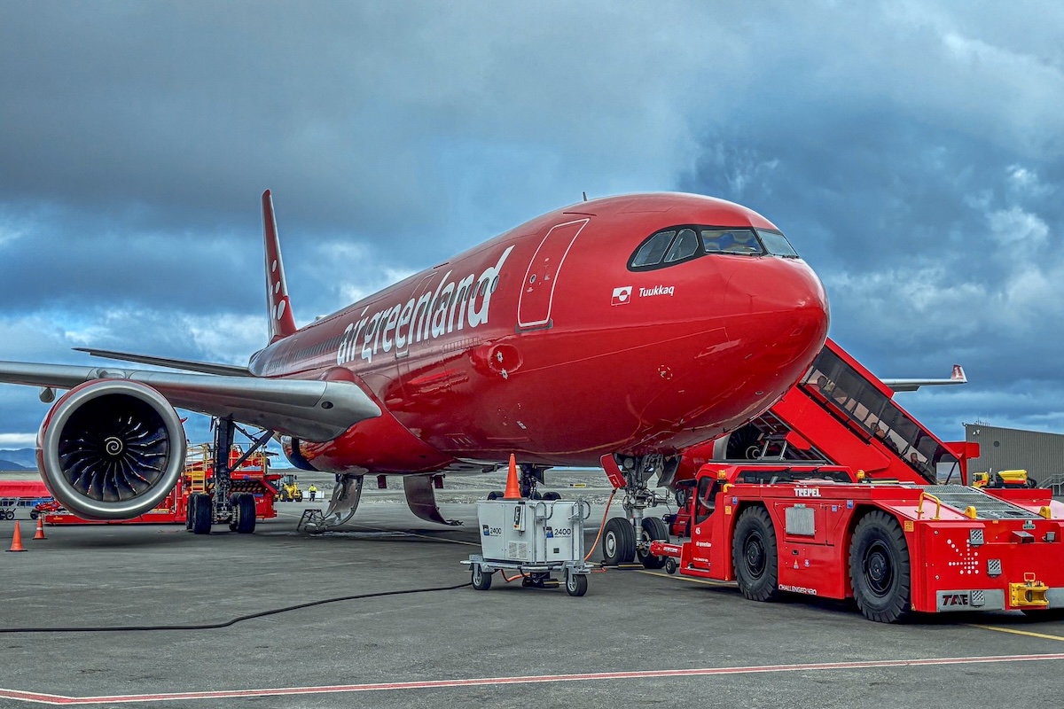 An A330-800neo in all-red Air Greenland livery at an airport gate under a grey sky. A red tug is connected to the front landing gear. 