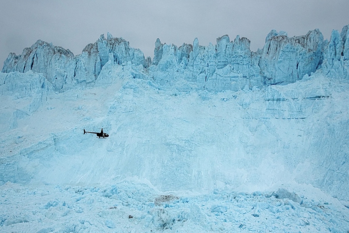 A dark-coloured helicopter flies along the terminal face of a massive, craggy glacier that towers above it. The sky above is uniform, medium grey. 