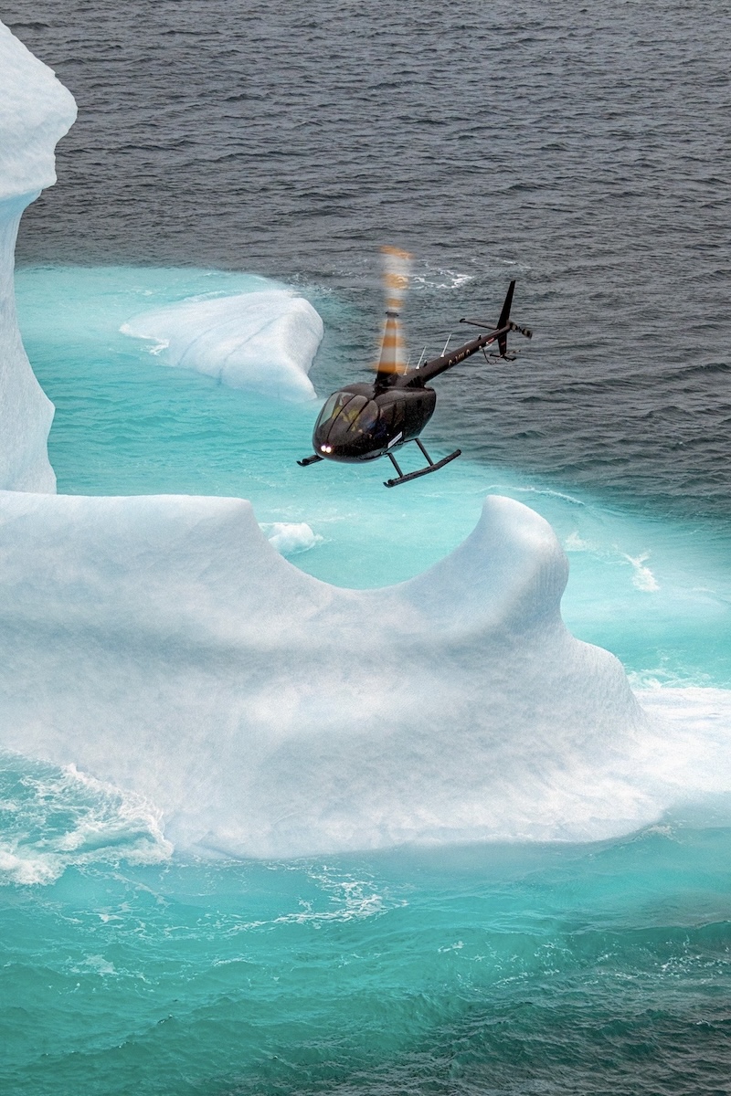 A helicopter hovers over an iceberg on a calm, grey sea. The iceberg above the water is white whereas the ice below is teal blue.
