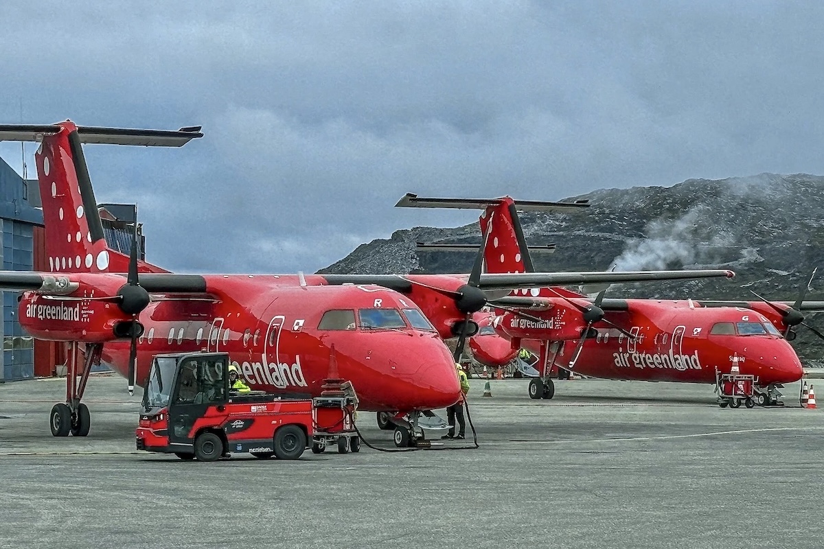 Two De Havilland Dash 8 type aircraft are parked adjacent to a green hangar. Both are painted similarly; that is, the all-red livery of Air Greenland. 