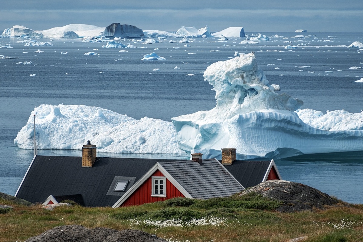 The roof of a house beyond a field of scrubby grass and wild flowers. Beyond, there is a calm sea with a large, jagged iceberg. 