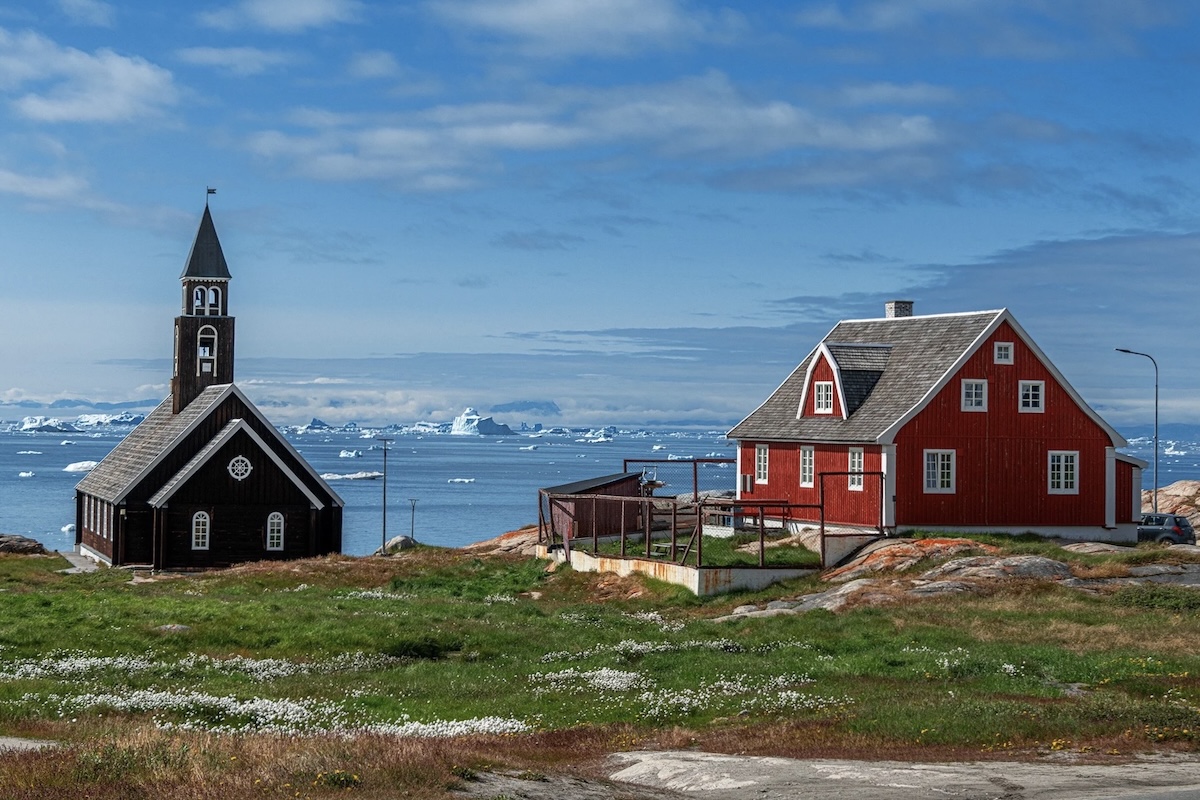 A church and a large red house are set in a field of green grass and wild flowers with a blue sea beyond. 