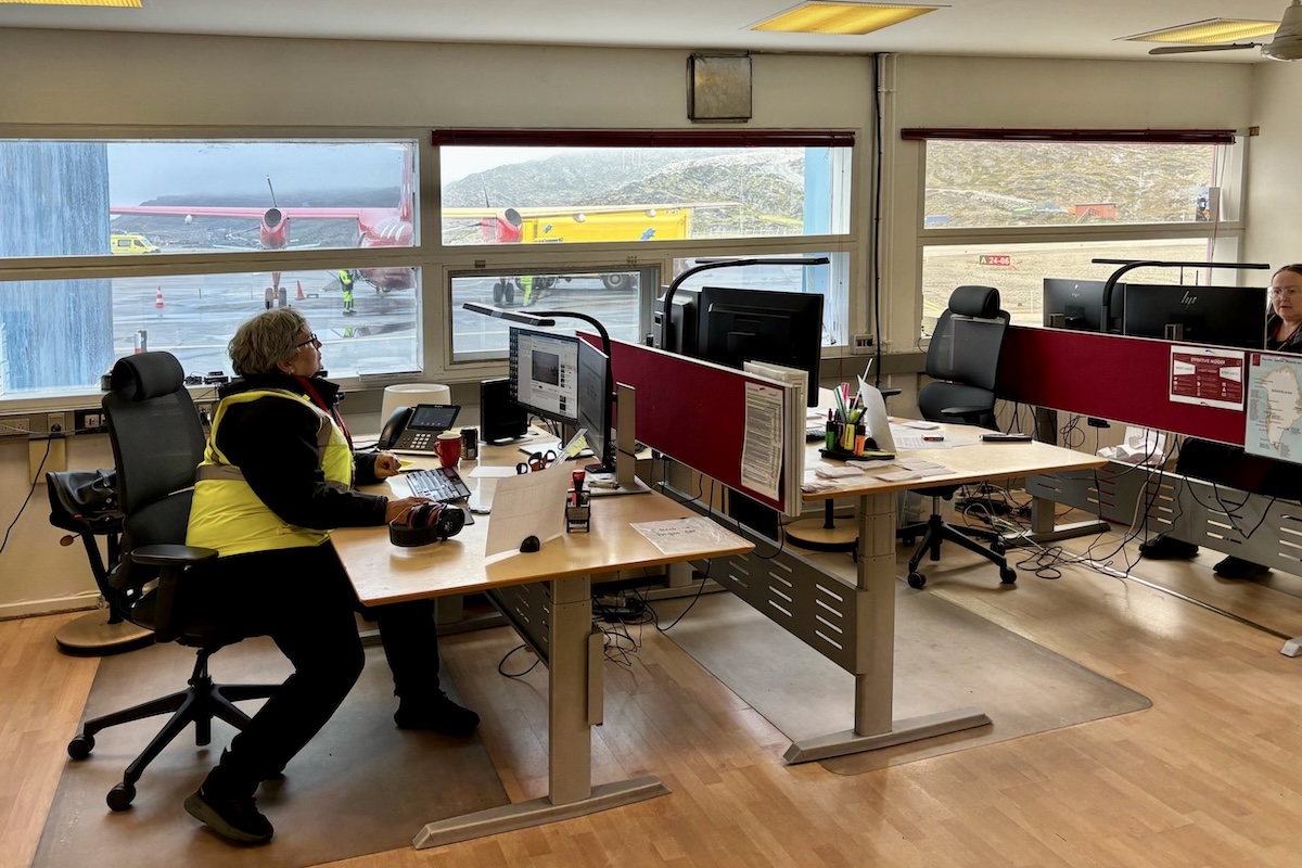 A room with closely spaced desks with computer monitors, with people sitting at two of them. A red aircraft is visible through large windows. 