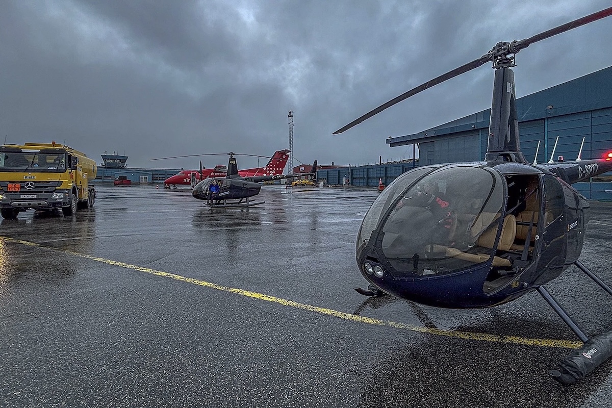 A rainy airport ramp with two dark-coloured helicopters and a red Dash 8, along with a yellow fuel truck, parked next to a green hangar. 