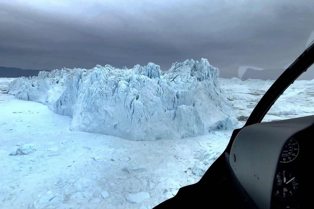 A pilot's perspective of a greenish, craggy iceberg floating amidst pack ice and smaller icebergs, along with the instrument panel of their helicopter. 