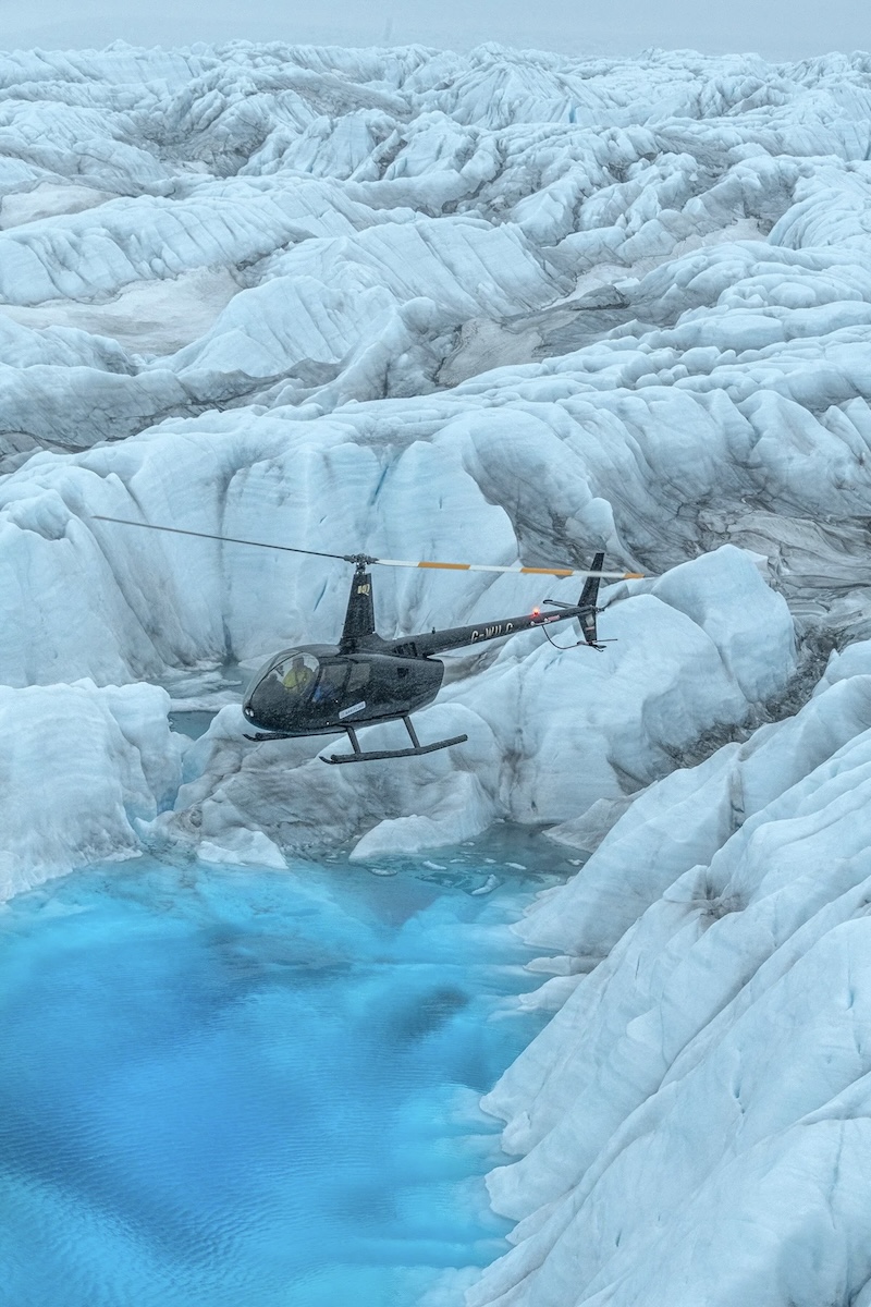 A dark-coloured helicopter hovers over a pool of meltwater set amongst a craggy glacier. The helicopter's blades are painted with red and white stripes.
