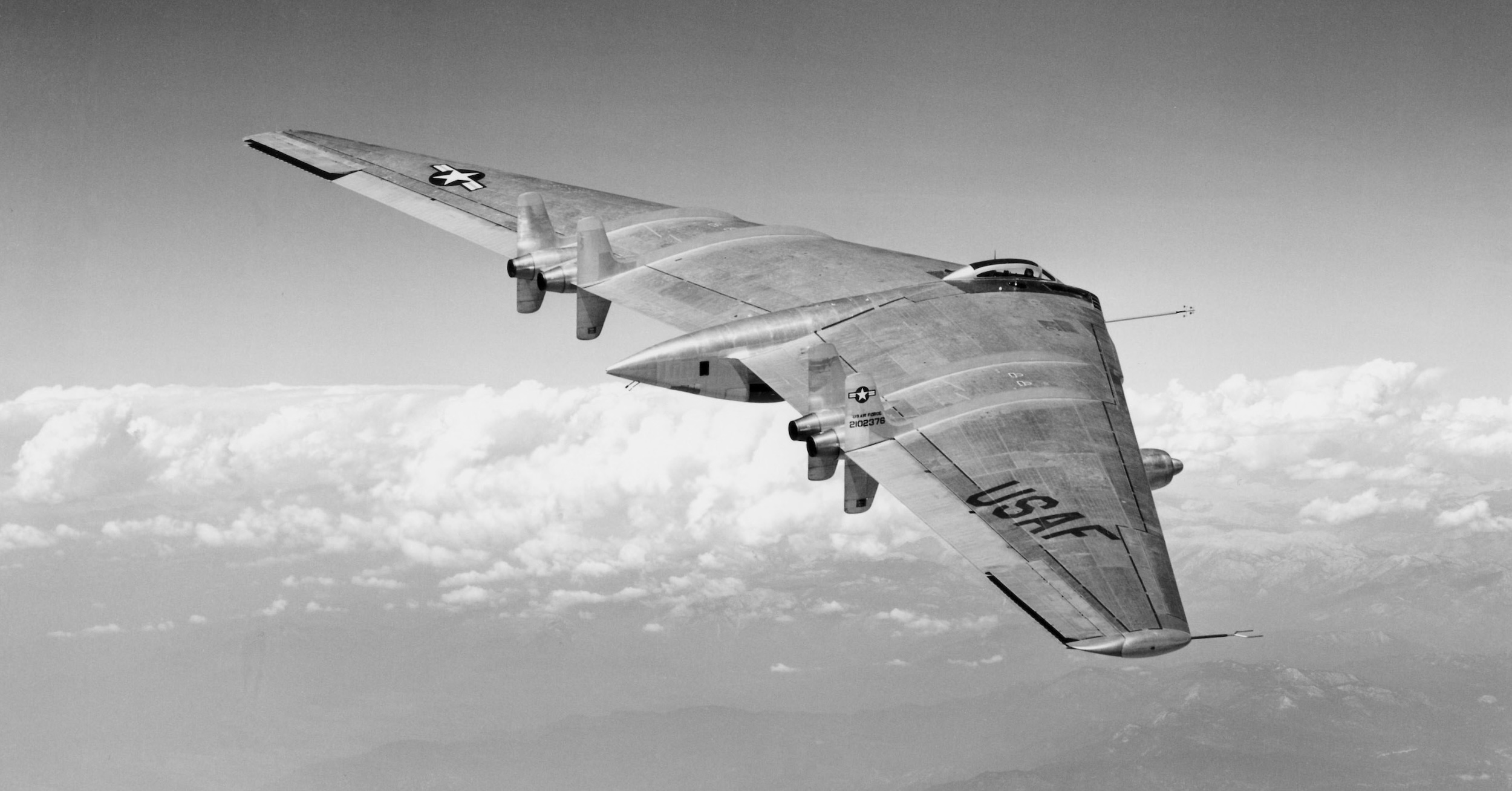 A black and white image of Jack Northrop's YB-49 flying wing design flying in a clear sky. Below, cumulus clouds are rising from the hilly, rolling terrain below. The aircraft is a simple wing with no fuselage or tail. There is a small, vestigial fuselage emerging from the centre trailing edge. Forward of this, there is a small bubble canopy where the pilot's helmet is visible. The aircraft appears to be bare metal with no paint coating. The camera is positioned to the right and slightly behind the aircraft, seemingly located in an aircraft flying in formation. The view of the aircraft is from the right wingtip and then along the wing's length. At the trailing edge, there are outlets from four jet engines embedded in the wing. Above and below each outlet is a small vertical stabiliser. On the nearest of these, there is a US Air Force 'roundel', then the words « US AIR FORCE » below it, and below this, the number « 2102376 ». On the upper surface of the right wing, the letters « USAF » are painted. The US Air Force roundel is painted on the upper surface of the left wing. What appears to be a fifth jet engine is visible just forward of the leading edge on the right wing. Some sort of data collection probe emerges from the leading edge of the wing near the pilot's position. Original caption from source: « The first flight of the all-jet-powered YB-49 on October 21, 1947. » (📸 Air Force Flight Test Center History Office) (📸 Air Force Flight Test Center History Office) »