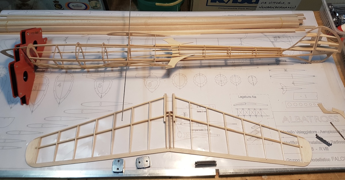 The framework of a model glider as it is being built. Nearest the camera is the framework for the horizontal stabiliser, which has a pleasing aftward sweep and taper from its root to its tips. Further away from the camera is the framework for the fuselage, which is running left and right in the frame. Beyond the fuselage is an array of wooden sticks seemingly used in the construction. A single wing rib is also visible. All of these elements are placed on a detailed, full-size plan for the glider. Original caption from source: « The Albatross in various stages of construction. » (📸 Paulo Rossi | Gruppo Falchi)