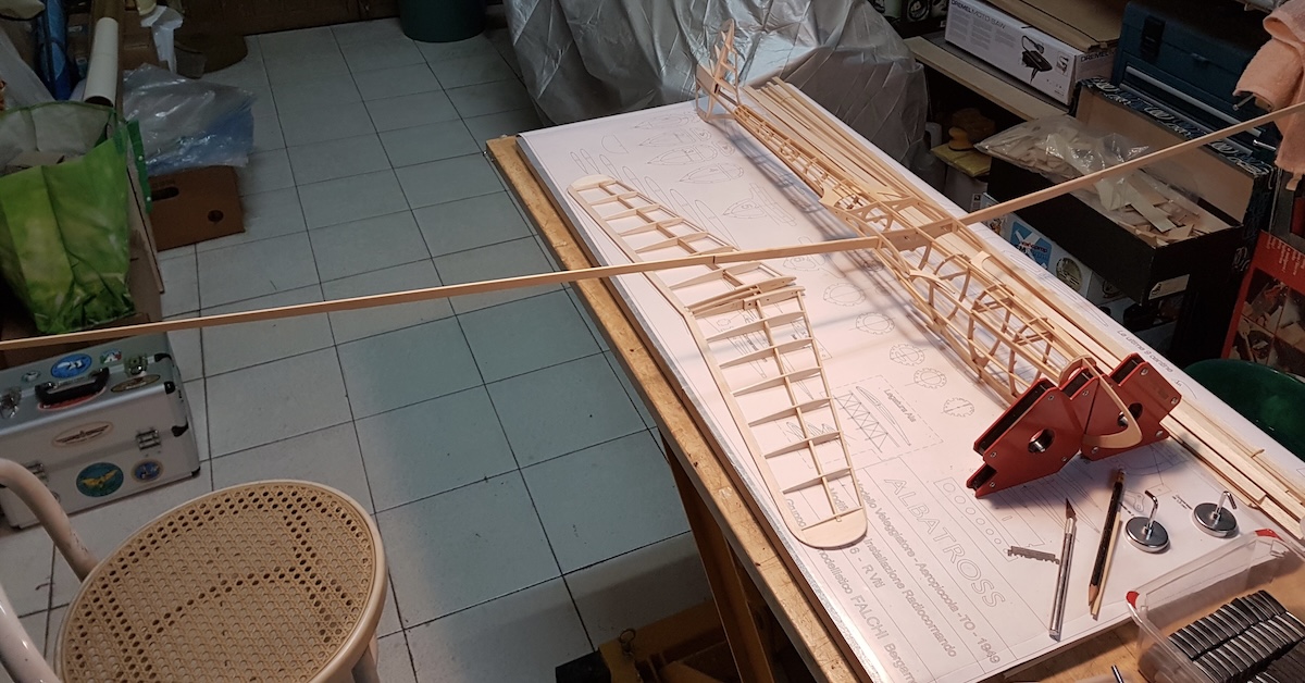 The framework of a model glider as it is being built. Nearest the camera is the framework for the horizontal stabiliser, which has a pleasing aftward sweep and taper from its root to its tips. To the right of the horizontal stabiliser is the framework for the fuselage, which is running toward and to the right of the camera position. The wing spar has been located in the fuselage in its final position, albeit only temporarily pending the rest of the wing construction. To the right of the fuselage is an array of wooden sticks seemingly used in the construction. All of these elements are placed on a detailed, full-size plan for the glider, which, in turn, is on a wooden workbench. There is a cane-covered chair to the left of the table. A tile floor and other workshop miscellany are also visible. Original caption from source: « The Albatross in various stages of construction. » (📸 Paulo Rossi | Gruppo Falchi)