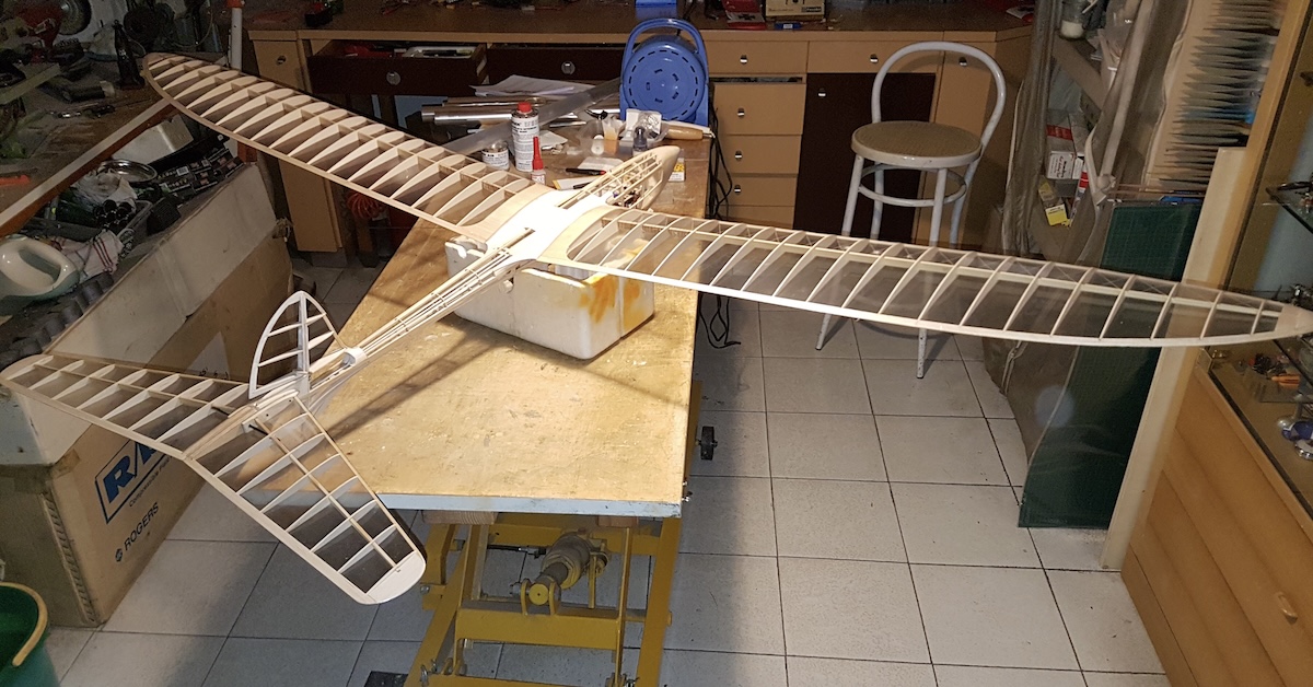 The seemingly complete framework for a model glider. It is not yet covered, so the full detail of the internal structure is clearly visible. The glider is resting on a small foam box, which in turn is resting on a wooden table. The aircraft is pointing away from the camera and to the right in the frame. The wing tips are pleasingly rounded, and the horizontal stabiliser tapers from root to tip. The vertical stabiliser is rounded in a manner that echoes the curves of the wing. The fuselage is constructed with formers oriented laterally, combined with thin stringers running fore and aft. Various shop tools and fixtures are also visible in the frame, as is another workbench and a chest of drawers beyond the workbench on which the glider is resting. Original caption from source: « The Albatross in various stages of construction. » (📸 Paulo Rossi | Gruppo Falchi)