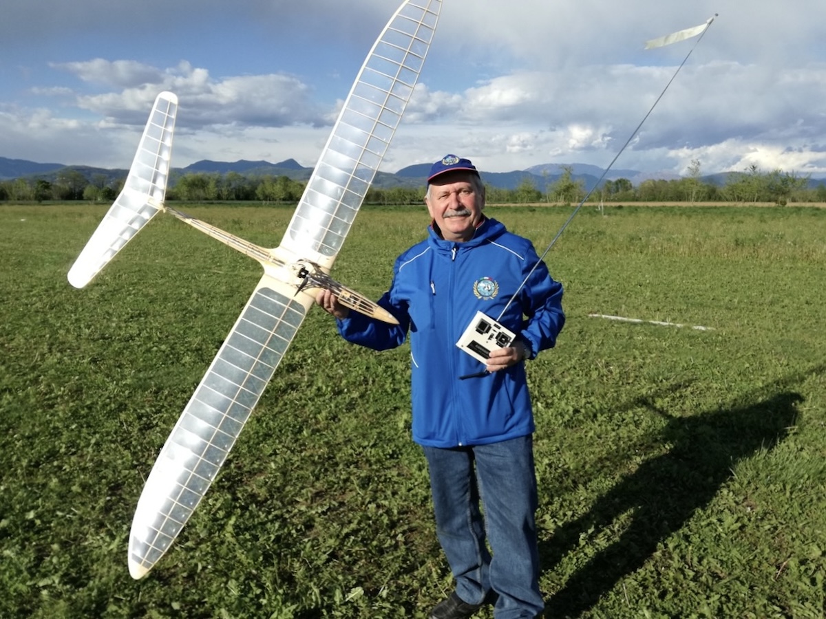 A middle-aged man with a grey moustache holds a radio-controlled model glider in his right hand while posing and smiling for the camera. The framework for the glider is clearly visible through its transparent skin. He holds in his right hand the transmitter, used to control the aircraft while in flight. He is wearing a blue jacket of light construction and is also wearing jeans and a blue hat. He is standing on what appears to be a  closely mown green field. Rolling hills are visible in the background. The subject of the photo is casting a sharp, dark shadow, indicating that the sun is shining. Original caption from source: « Gruppo Falchi members who participated in the club build of the Aeropiccola Albatross. » (📸 Paulo Rossi | Gruppo Falchi)