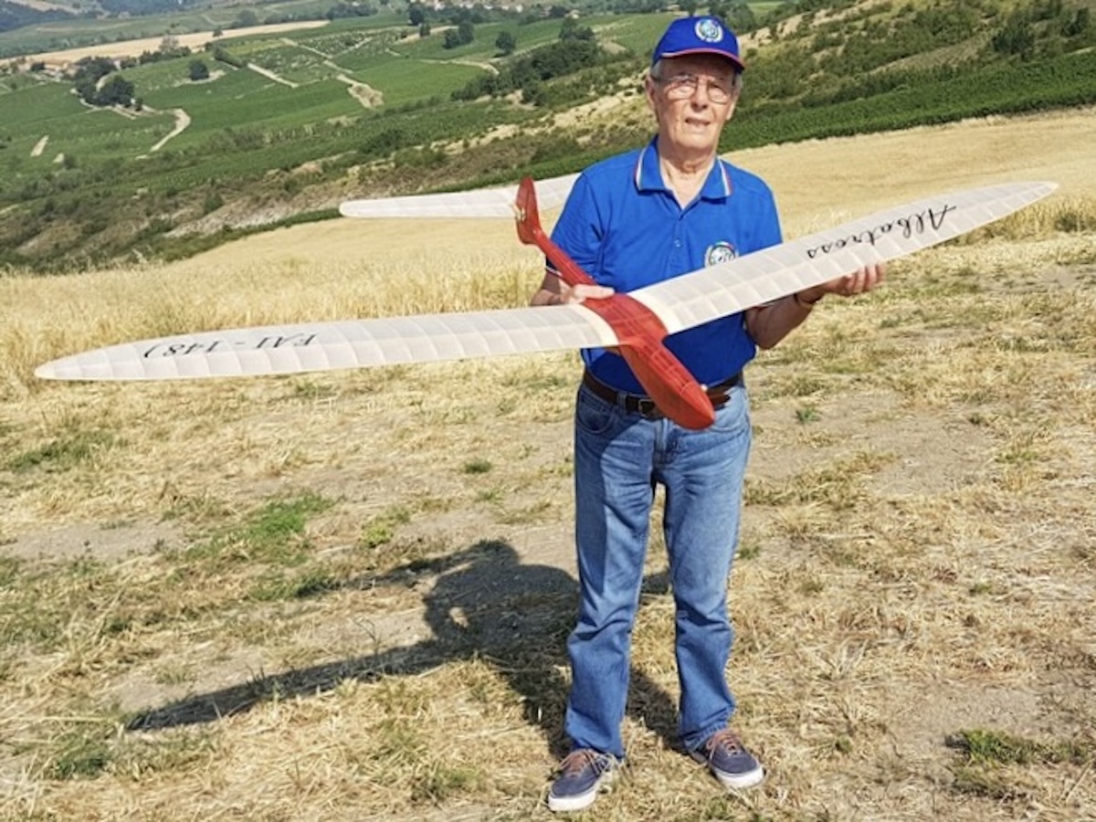 An older man holds a radio-controlled model glider while posing and smiling for the camera. The wing is slightly translucent white, and the fuselage of the glider is red. He is wearing a blue, open-collar shirt and jeans. He is also wearing prescription glasses. He is standing on a sloping field with longish, yellow grass. Rolling hills of varying gold and green sections are visible in the background. The subject of the photo is casting a sharp, dark shadow, indicating that the sun is shining. Original caption from source: « Gruppo Falchi members who participated in the club build of the Aeropiccola Albatross. » (📸 Paulo Rossi | Gruppo Falchi)