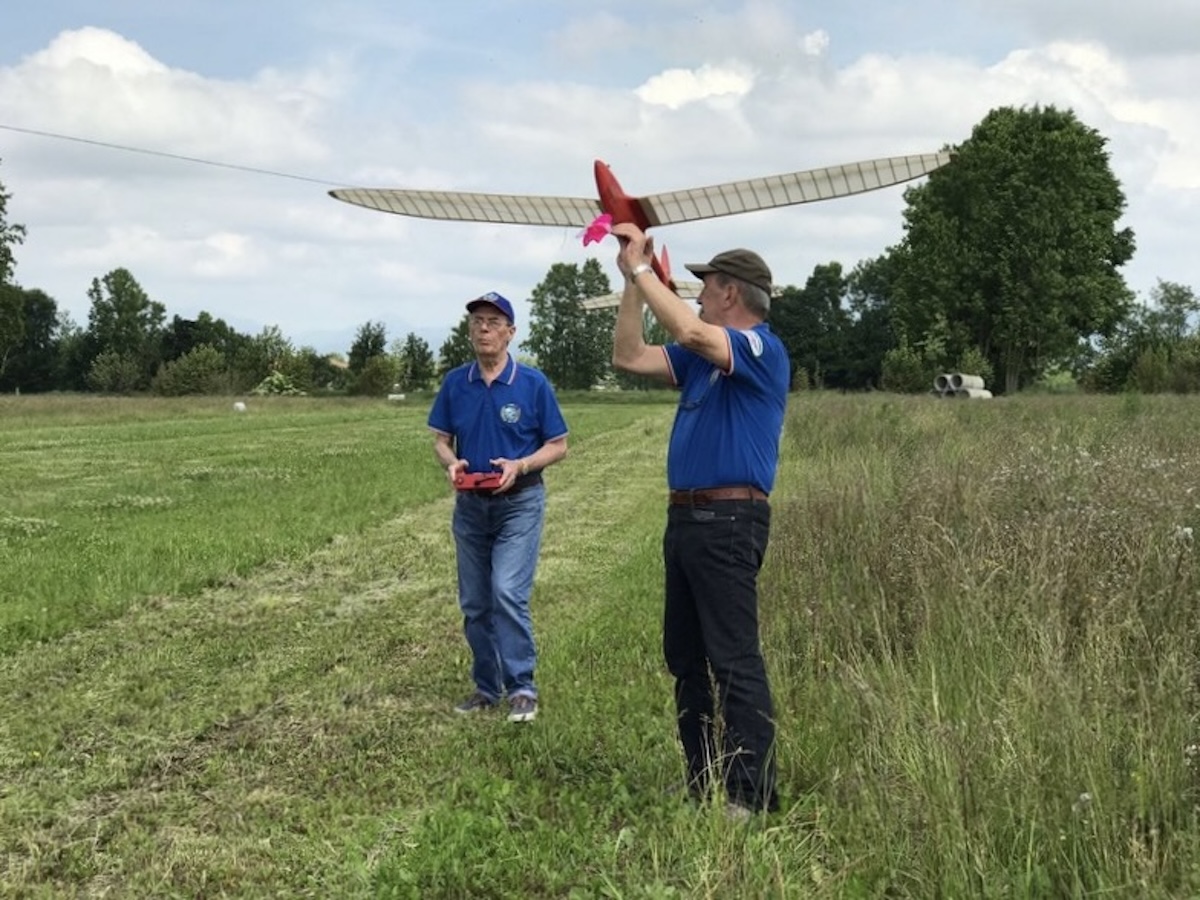 Two middle-aged men prepare a radio-controlled model glider for launch on a towline. One of the men is holding the glider aloft, as if to launch it, and the second man stands to the left in the frame and is holding the transmitter used to control the aircraft while it is in flight. Once launched, it appears as though the aircraft will be flying upwards, toward and slightly to the left of the camera position. The wing is slightly translucent white, and the fuselage of the glider is red. Both men are wearing a blue, open-collar shirt and jeans. The man holding the glider is looking at the man with the transmitter, and they appear to be engaged in a conversation. They are standing on a field of mown grass adjacent to an area with longer grass. A line of trees is visible in the background. The subjects of the photo are casting an indistinct shadow, indicating an overcast day. Original caption from source: « Gruppo Falchi members who participated in the club build of the Aeropiccola Albatross. » (📸 Paulo Rossi | Gruppo Falchi)