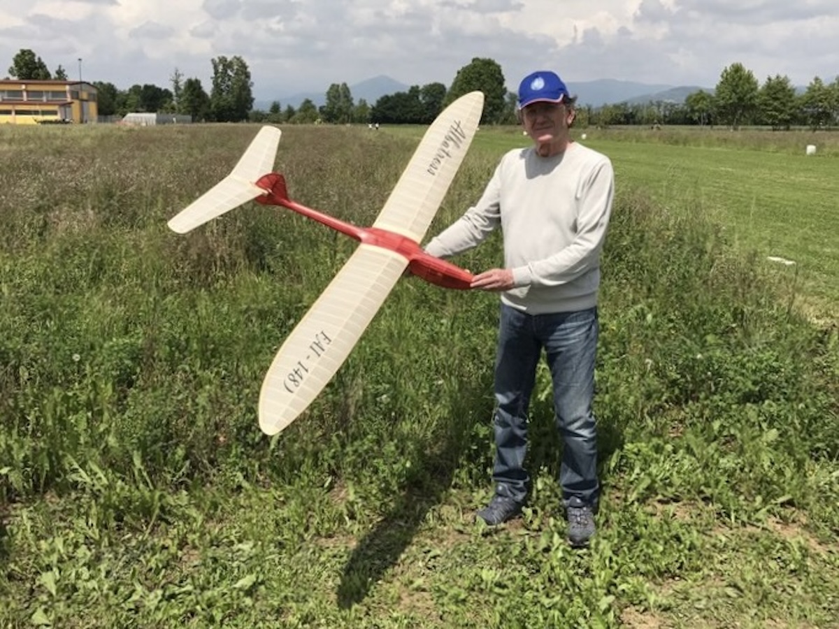 A middle-aged man holds a radio-controlled model glider to his right while posing and smiling for the camera. The wing is slightly translucent white, and the fuselage of the glider is red. The word « Albatross » is written in black script on the left wing. The words « FAI — 1481 » are written on the right wing in a similar script. He is wearing a cream-coloured sweater and jeans. He is also wearing a medium blue baseball hat with a light blue round logo. He is standing in a field of long, green grass which is adjacent to a closely mown strip. A line of trees and a building are visible in the middle distance. Beyond this, rolling hills can be seen in the far distance. The subject of the photo is casting a fairly sharp, dark shadow, indicating that the sun is shining although there is a cloudy sky visible in the photo. Original caption from source: « Gruppo Falchi members who participated in the club build of the Aeropiccola Albatross. » (📸 Paulo Rossi | Gruppo Falchi)