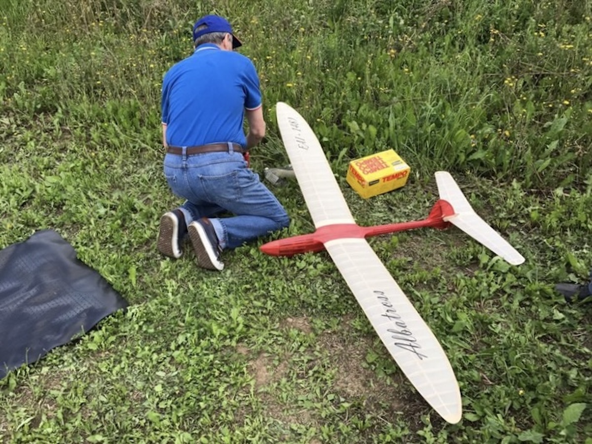 A man of indeterminate age kneels next to  a radio-controlled model glider, on the ground to his right, and has his back to the camera. The wing is slightly translucent white, and the fuselage of the glider is red. The word « Albatross » is written in black script on the left wing. The words « FAI — 1481 » are written on the right wing in a similar script. He is wearing a blue golf shirt and jeans. He is also wearing a medium blue baseball hat. There is a small yellow box with red writing adjacent to the glider, which might contain parts and pieces related to the work being done on the glider. Original caption from source: « Gruppo Falchi members who participated in the club build of the Aeropiccola Albatross. » (📸 Paulo Rossi | Gruppo Falchi)