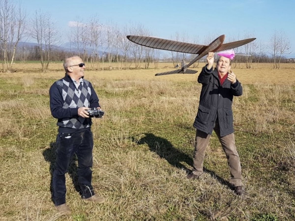 An older man holds a radio-controlled model glider to his right while posing and smiling for the camera. The wing is mostly slightly translucent white, and the fuselage of the glider is gold, as are the adjacent wing roots. He is wearing a blue argyll sweater and jeans. He is also wearing sunglasses. He is standing in a field of closely mown grass with a white dotted line, similar to what would be found on a road or runway, running fore and aft in the photo. There are smaller white markers at the edge of the mown strip, in lines parallel to the main line down the centre of the mown strip. A line of trees is visible in the middle distance. Beyond this, rolling hills can be seen in the far distance. The subject of the photo is casting a fairly sharp, dark shadow, indicating that the sun is shining, although. The sky above is clear blue dotted with a few puffy clouds. Original caption from source: « Gruppo Falchi members who participated in the club build of the Aeropiccola Albatross. » (📸 Paulo Rossi | Gruppo Falchi)