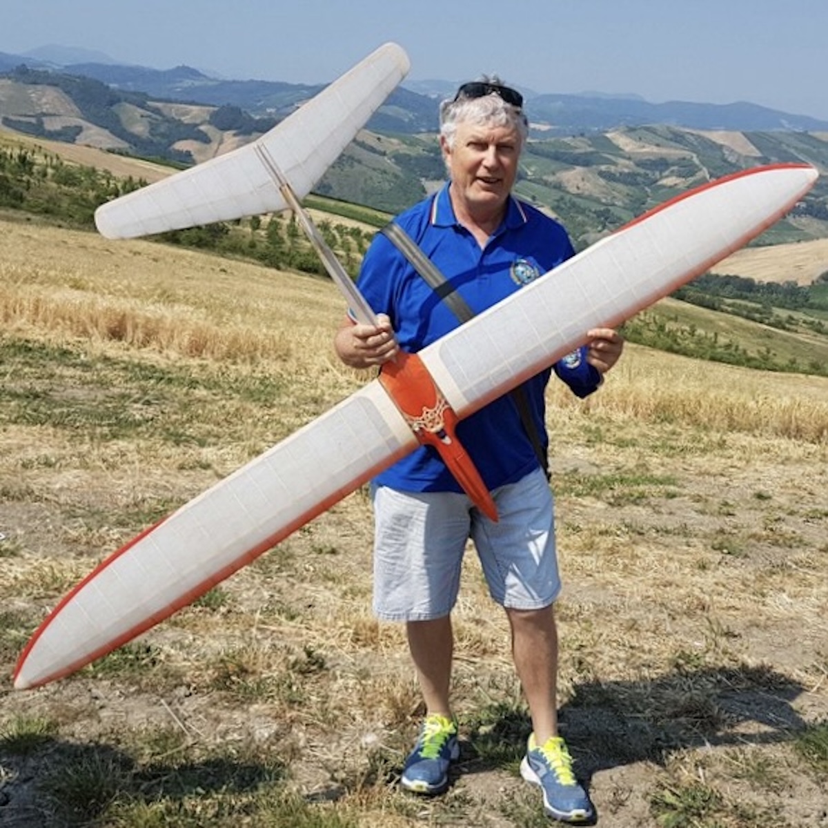 Two middle-aged men prepare a radio-controlled model glider for launch on a towline. One of the men is holding the glider aloft, as if to launch it, and the second man stands to the right in the frame with his back to the camera. Once launched, it appears as though the aircraft will be flying upwards, toward and slightly to the left of the camera position. The wing is slightly translucent white, and the fuselage of the glider is red. Both men are wearing a blue, open-collar shirt and jeans. They are standing on a field of mown grass adjacent to an area with longer grass. A line of trees is visible in the background. The subjects of the photo are casting an indistinct shadow, indicating an overcast day. Original caption from source: « Gruppo Falchi members who participated in the club build of the Aeropiccola Albatross. » (📸 Paulo Rossi | Gruppo Falchi)