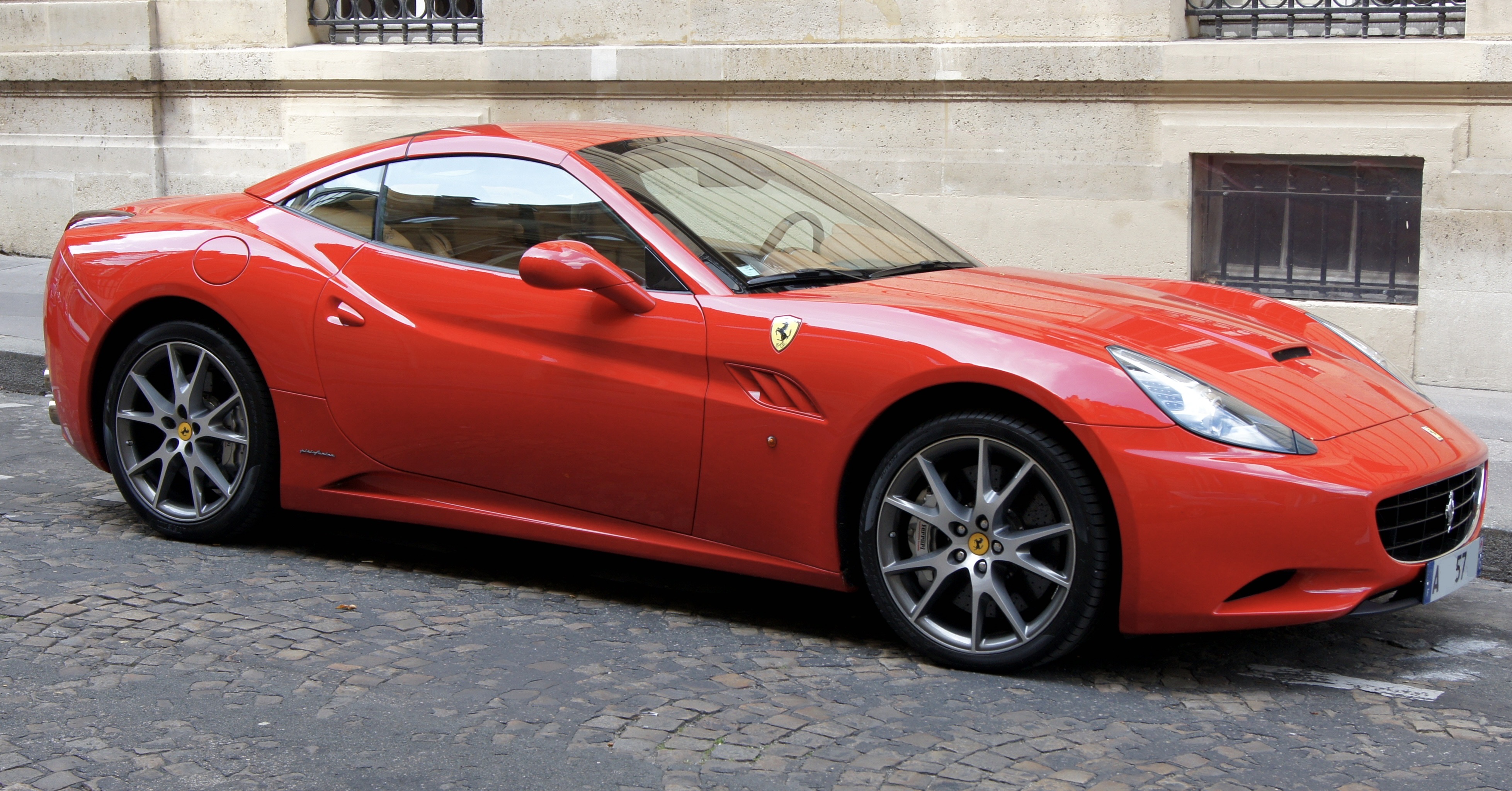 A 'Ferrari Red' Ferrari California is parked on a cobbled street. A building made of light-coloured stone is visible in the background. The car is oriented so the front is pointing toward the right side of the frame. The yellow Scuderia Ferrari shield appears on the fender just aft of the right front wheel. The license plate is just visible, with the letters « A 57 ». Original caption from source: « Ferrari California in a street of Paris. » (📸 Jebulon | Wikimedia | public domain)